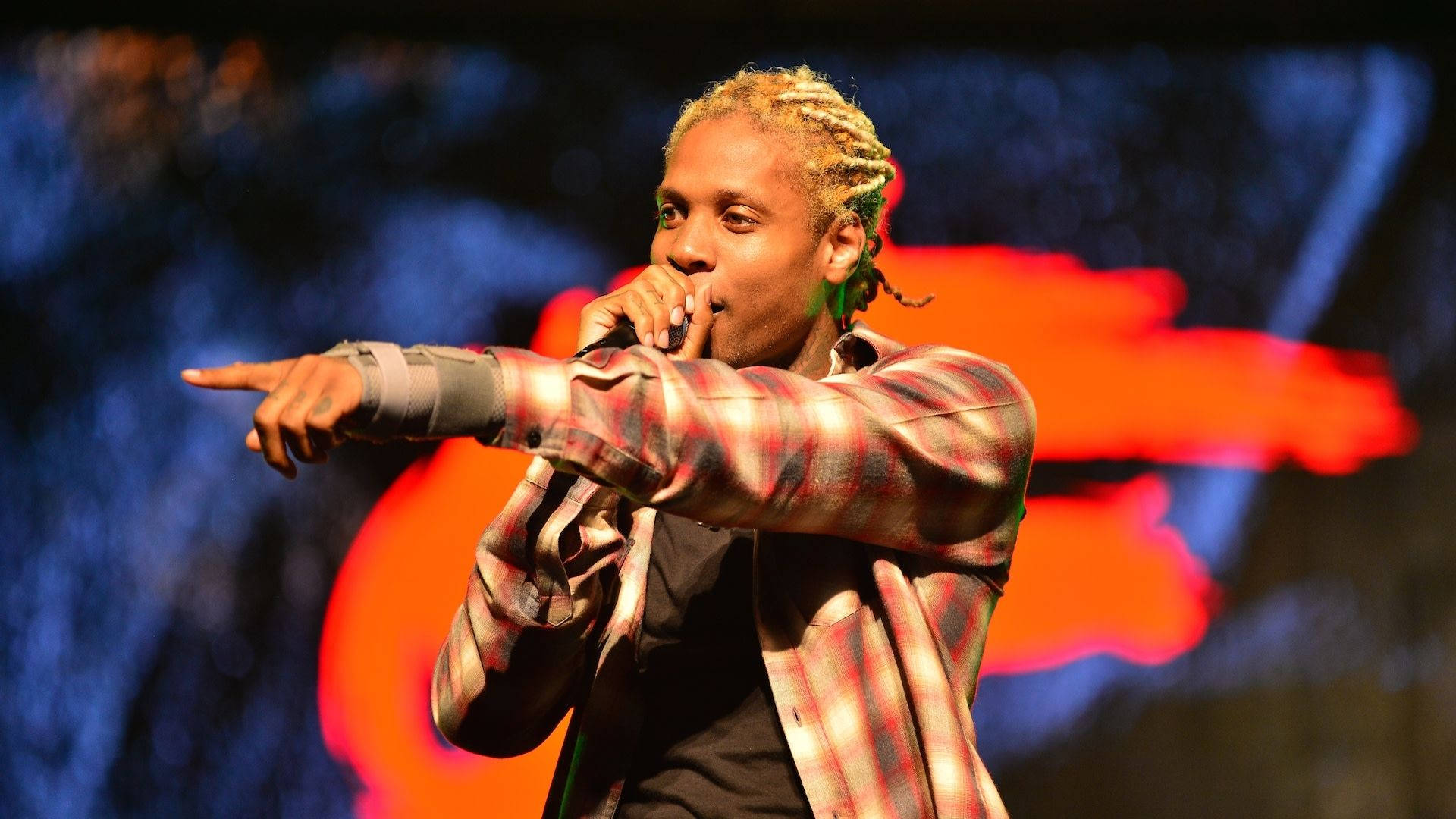 Lil Durk 與粉絲互動指揮 壁紙 — 一位辮子頭髮的男性表演者穿著格仔襯衫，在充滿活力的舞台燈光下指向前方，同時對著咪高峰唱歌。