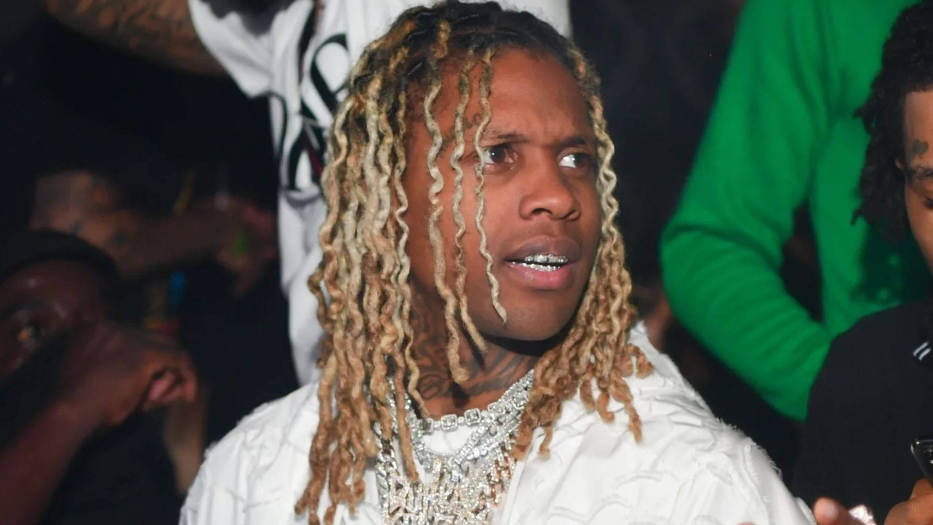 Lil Durk 與粉絲同框 壁紙 — 一名留著金色長髮辮、戴著鑽石鏈條的男子，在昏暗的人群中向上仰望。