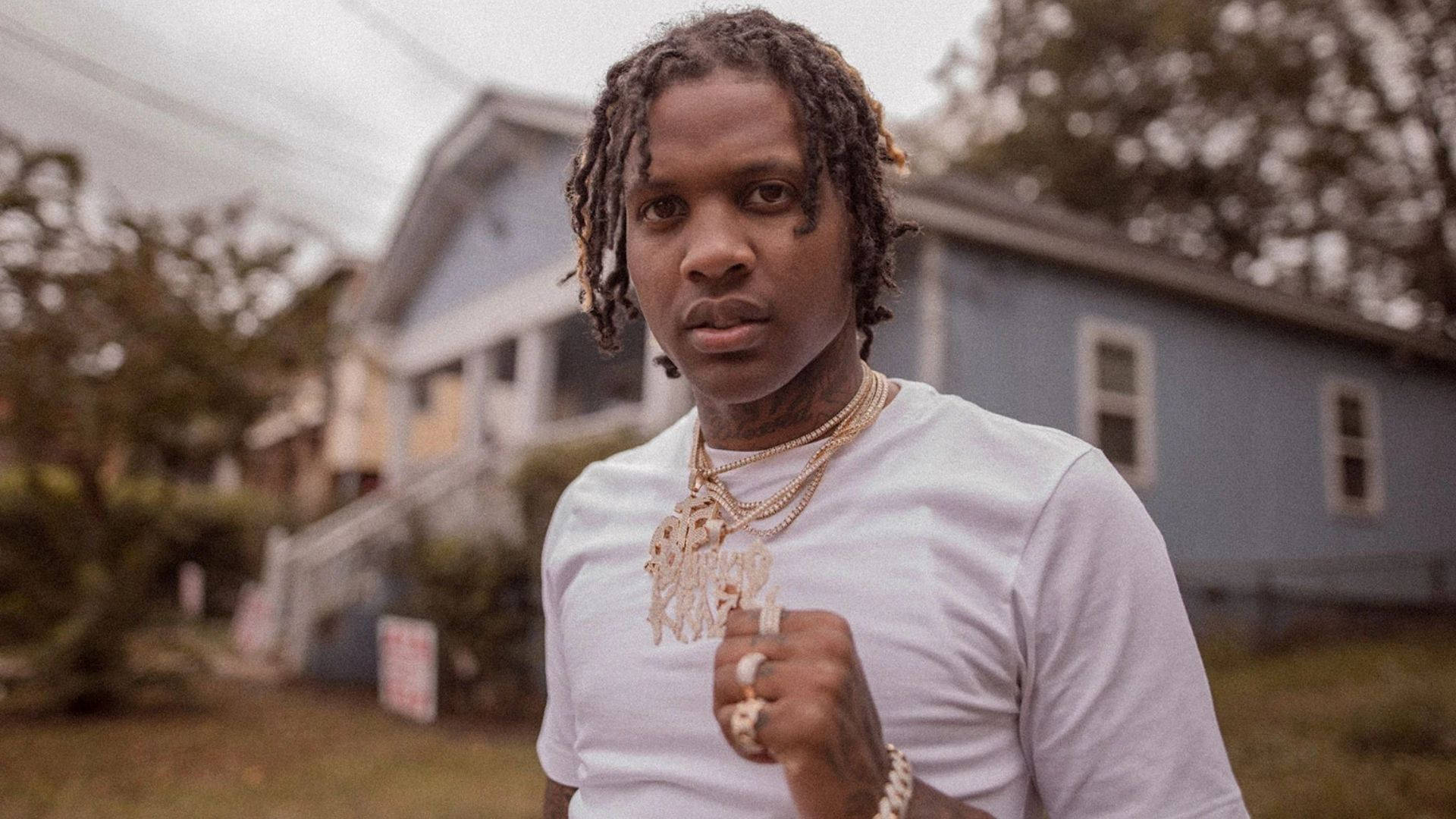 Lil Durk 舊屋前 壁紙 — 一位年輕男子梳著辮子、戴著金鏈，在陰天自信地站在藍色房子前。