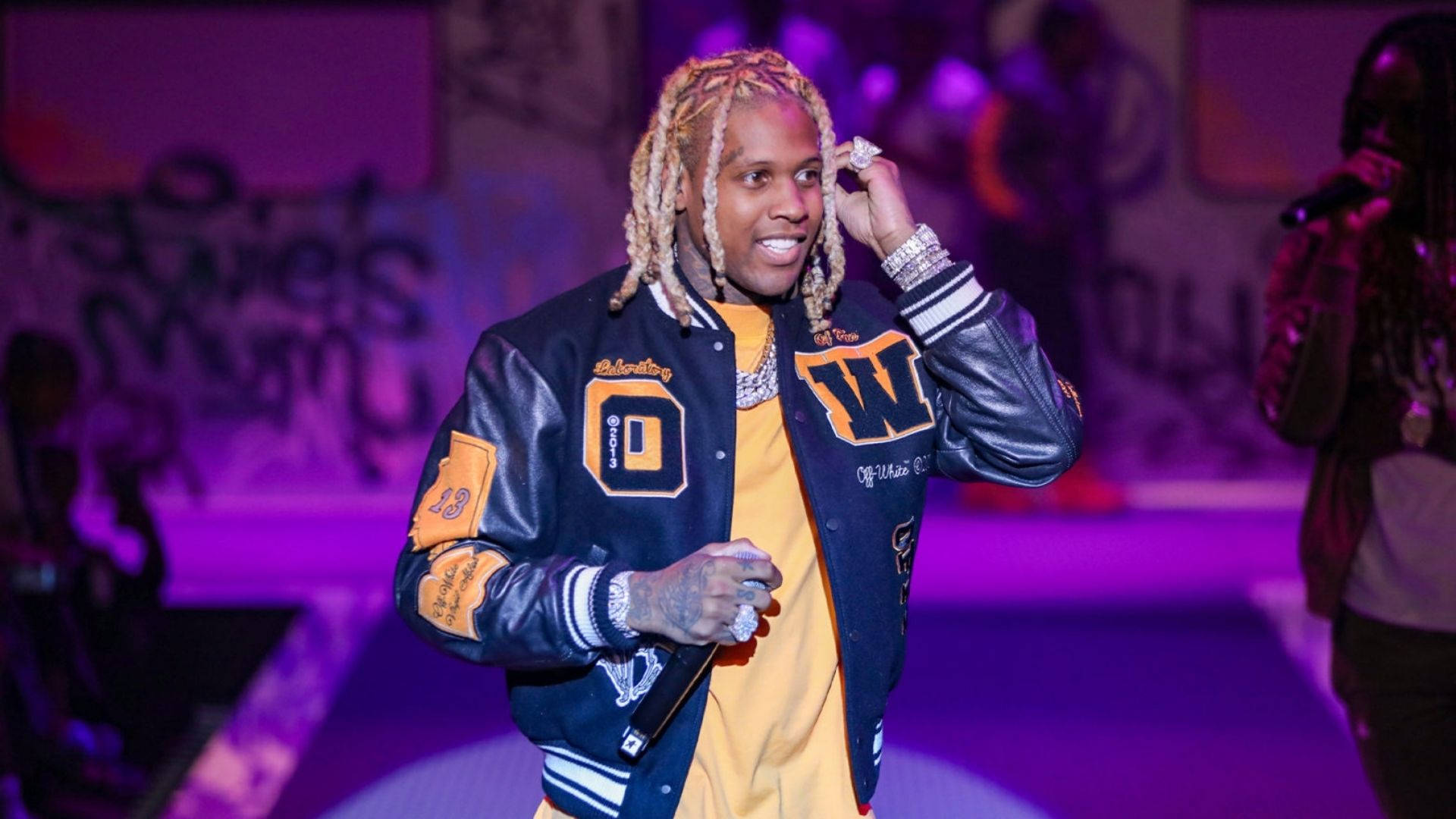 Lil Durk 舞台演出 壁紙 — 一位辮子頭髮的饒舌歌手在紫色燈光、塗鴉背景的舞台上微笑著調整耳朵。