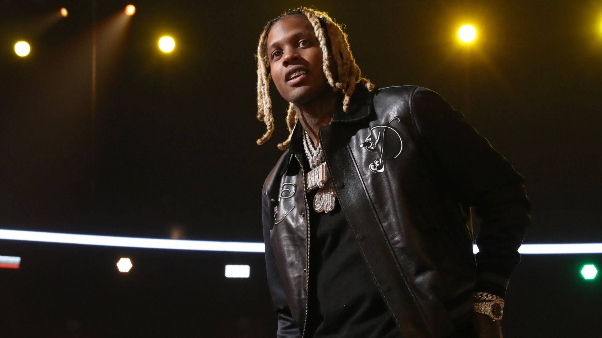 Lil Durk 舞台造型 壁紙 — 一位梳著辮子的說唱歌手自信地站在昏暗的舞台上，在溫暖的聚光燈下，身穿黑色皮夾克和戴著金鍊。