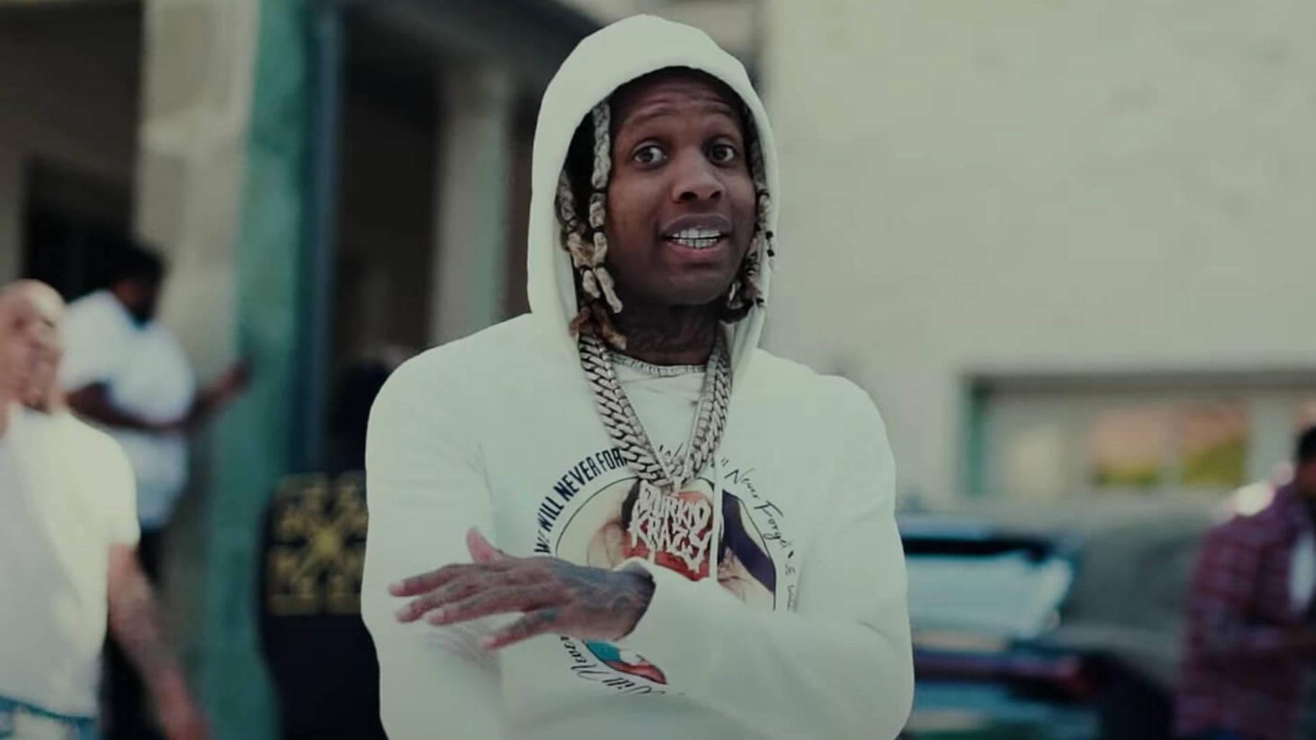 Lil Durk 街頭表演 壁紙 — 一名身穿白色連帽衫、辮子頭髮和粗鏈條的男子在戶外生動地做手勢。