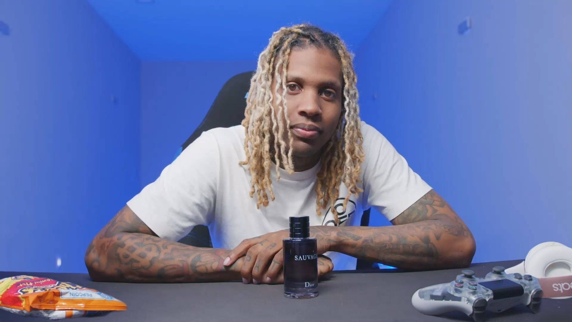 Lil Durk 遊戲房間 壁紙 — 一名擁有金色長髮辮和紋身的男子坐在藍色房間的書桌前，手持香水瓶，附近有一個遊戲控制器。