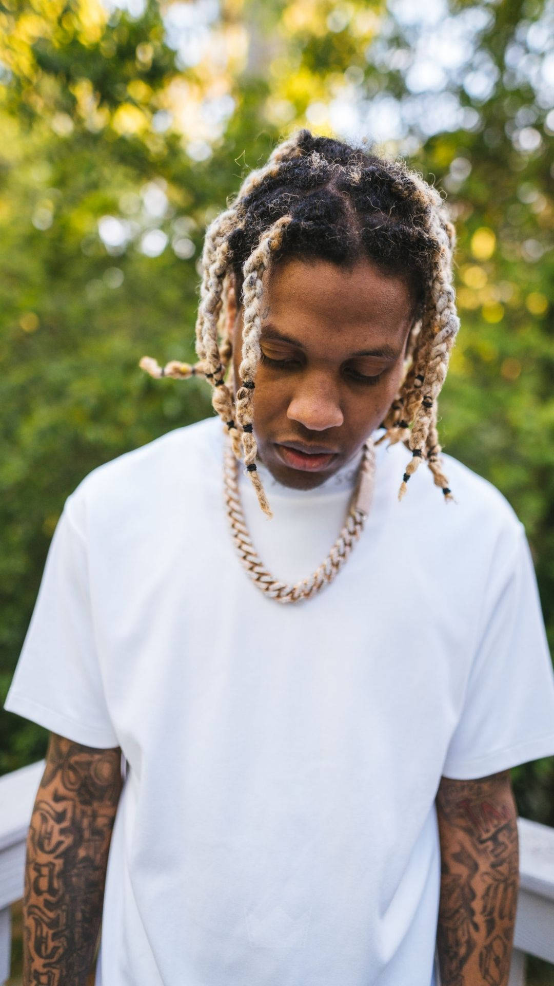 Lil Durk 金鏈造型 壁紙 — 一位辮髮紋身的年輕男子，穿著白色T恤和鏈條項鍊，在陽光普照的綠色背景下低頭凝視。