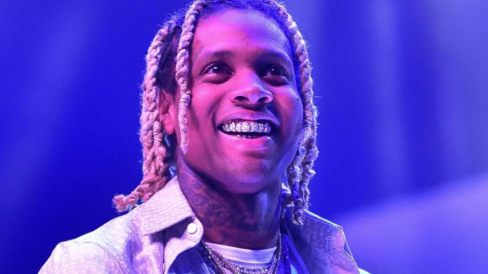 Lil Durk 開心微笑 壁紙 — 一個微笑的男人，梳著辮子頭，戴著鑽石牙套，在充滿活力的藍色舞台燈光下。