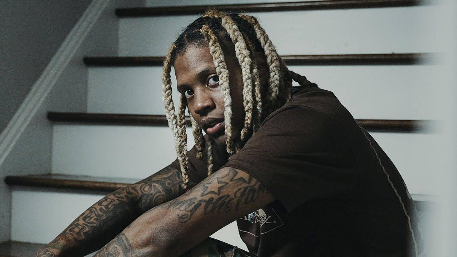 Lil Durk 霸氣神情 壁紙 — 一位年輕男子編著辮子、身上有紋身，坐在樓梯上，專注地凝視鏡頭。