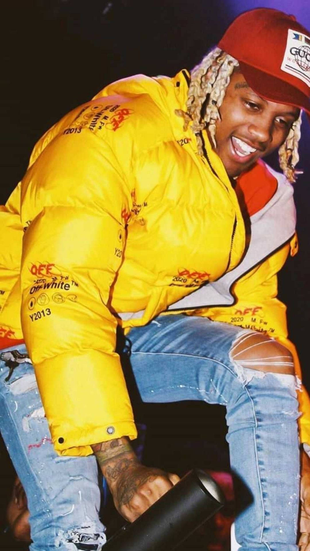 Lil Durk 黃色夾克造型 壁紙 — 一位微笑的男子，編著辮子，身穿亮黃色羽絨外套和破洞牛仔褲，在舞台上拿著麥克風。