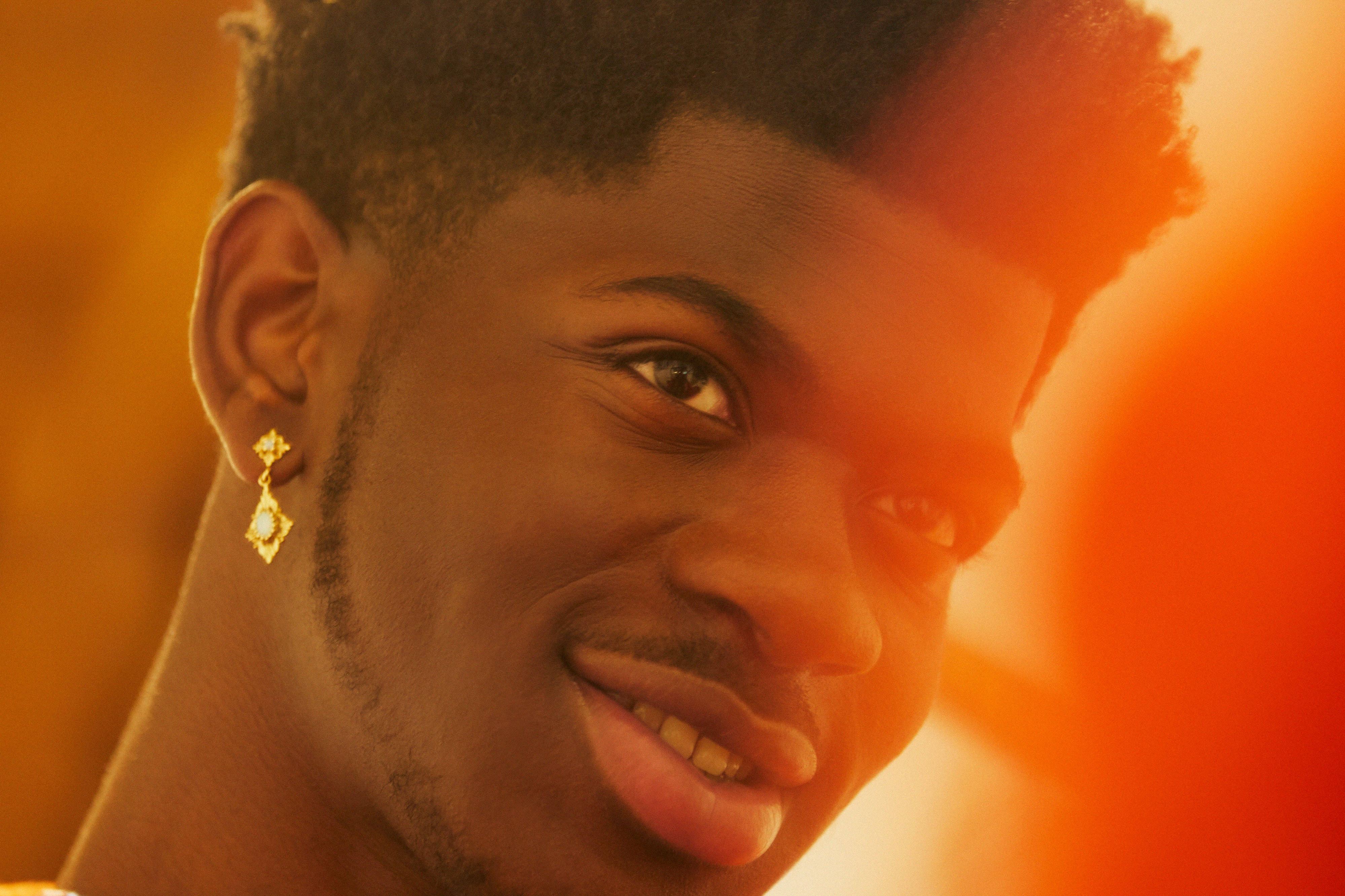 Lil Nas X GQ 杂誌拍攝 壁紙 — 一位年輕男子帶著溫暖的笑容和金耳環，在充滿活力的橙色日落背景下閃耀。