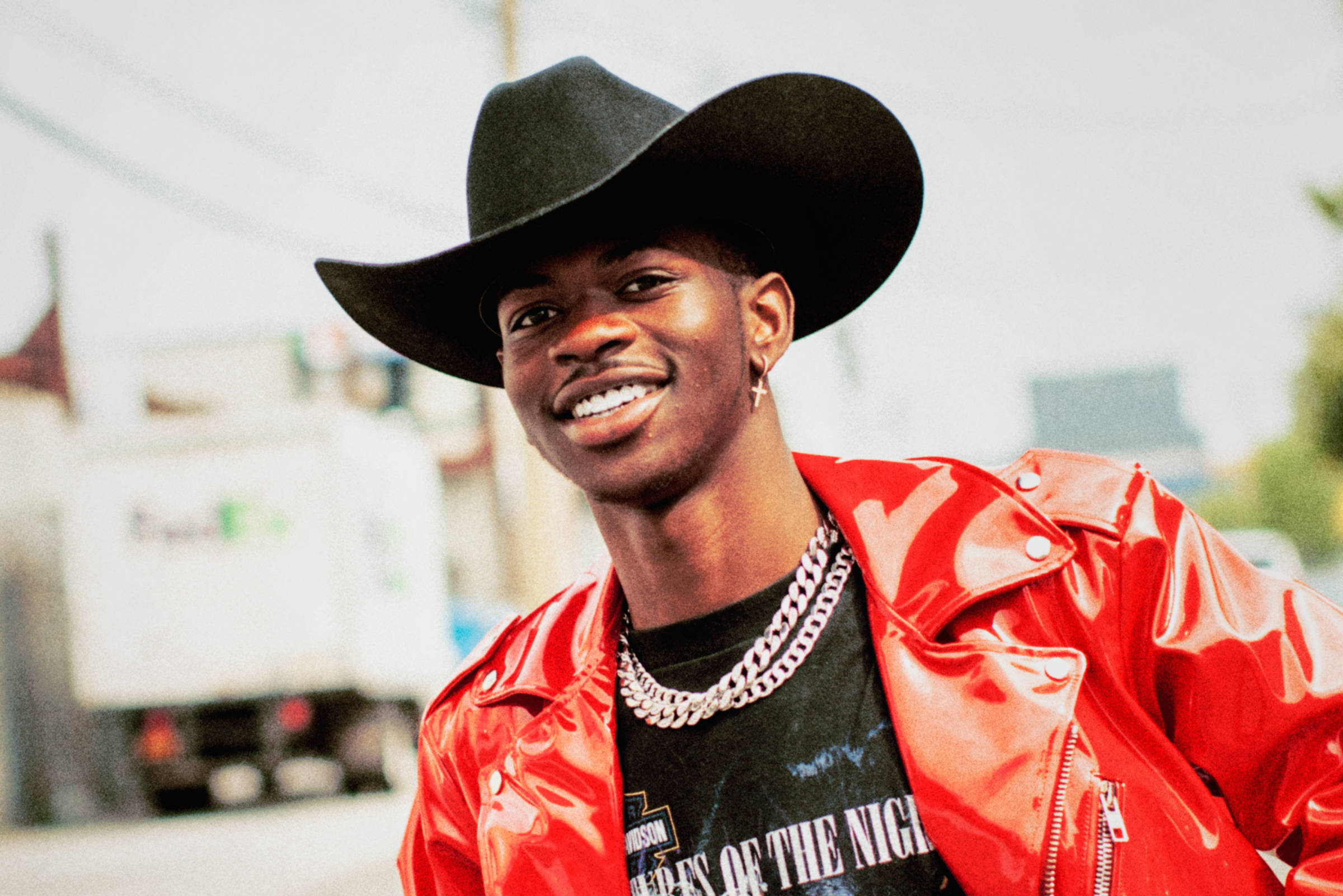 Lil Nas X Rolling Stone 杂誌拍攝 壁紙 — 一位戴著黑色牛仔帽、身穿閃亮紅色外套的年輕男子微笑著，自信地在城市街道上擺姿勢。