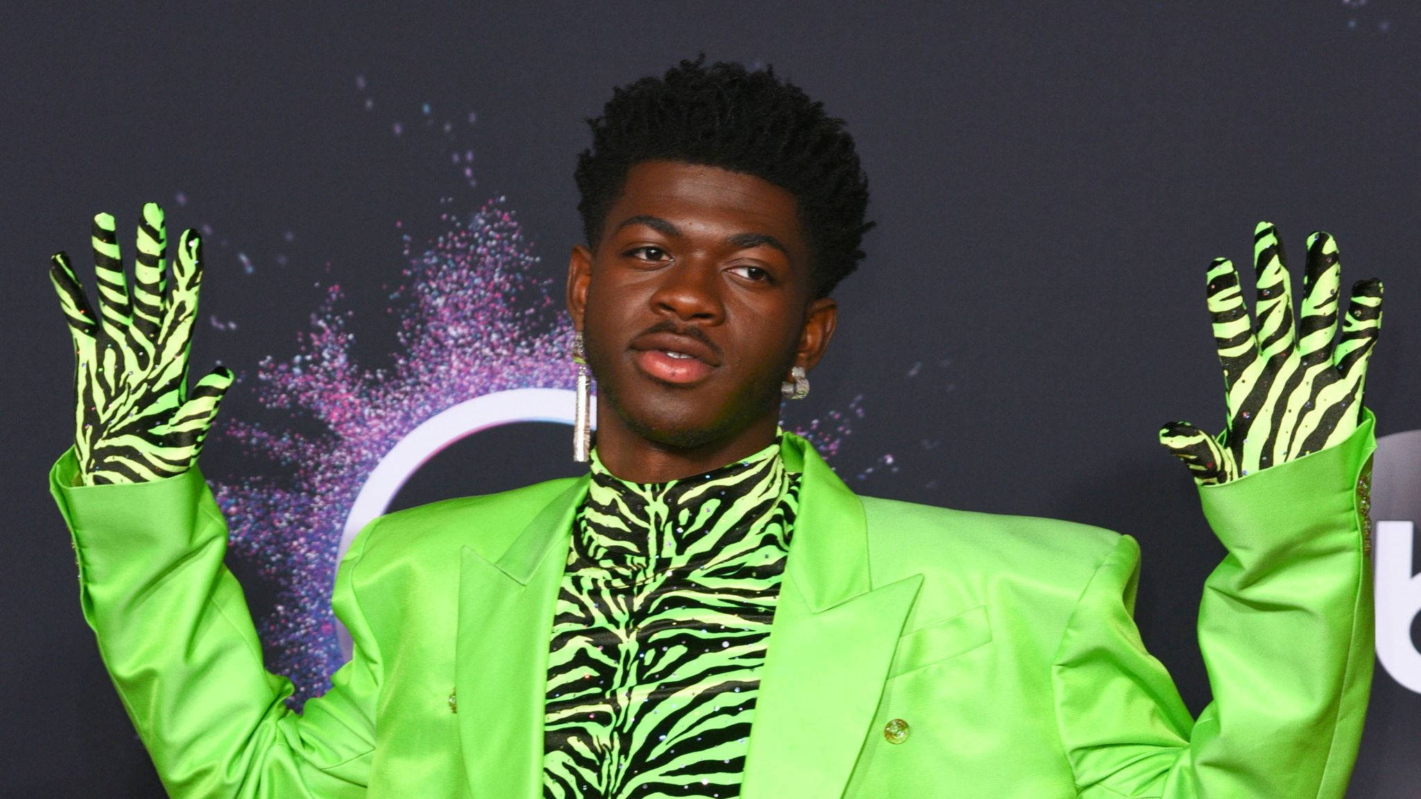 Lil Nas X 嘻哈歌手造型 壁紙 — 一名身穿亮綠色西裝和斑馬紋手套的男子，在帶有紫色閃粉的深色背景前舉起雙手擺姿勢。