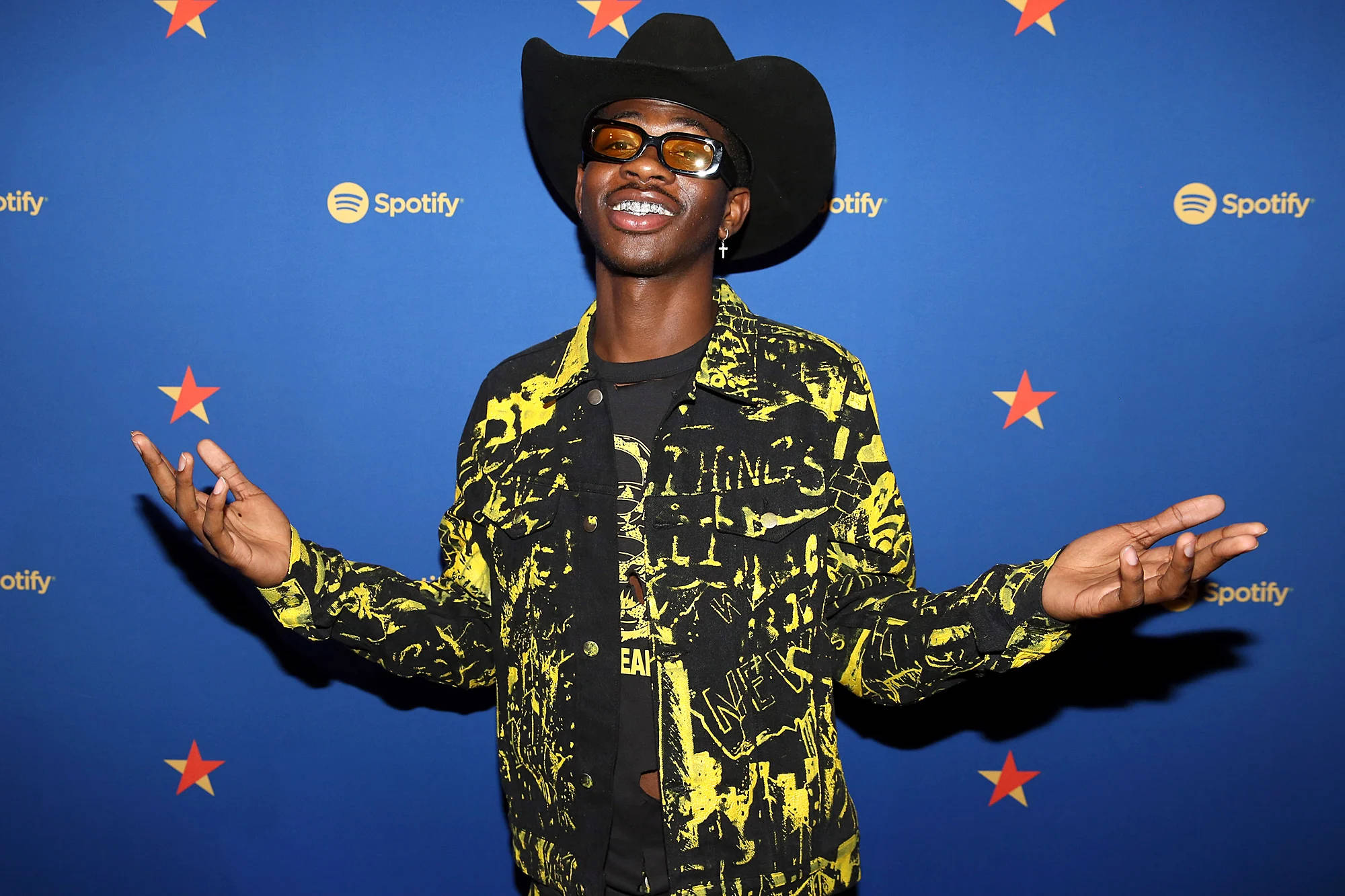 Lil Nas X 於 Spotify 活動 壁紙 — 一個微笑的男人，戴著黑色牛仔帽和黃色圖案外套，張開雙臂，以帶有紅色星星的藍色Spotify背景為襯托。