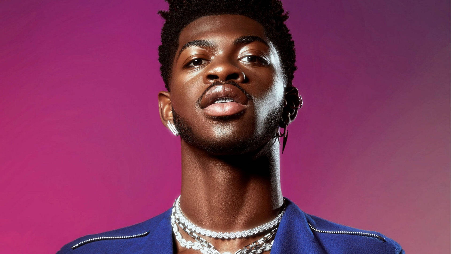 Lil Nas X 的 Montero Hill 壁紙 — 一位自信的年輕男子，梳著時尚髮型，戴著多層鏈條，在充滿活力的洋紅色漸變背景前直視鏡頭。