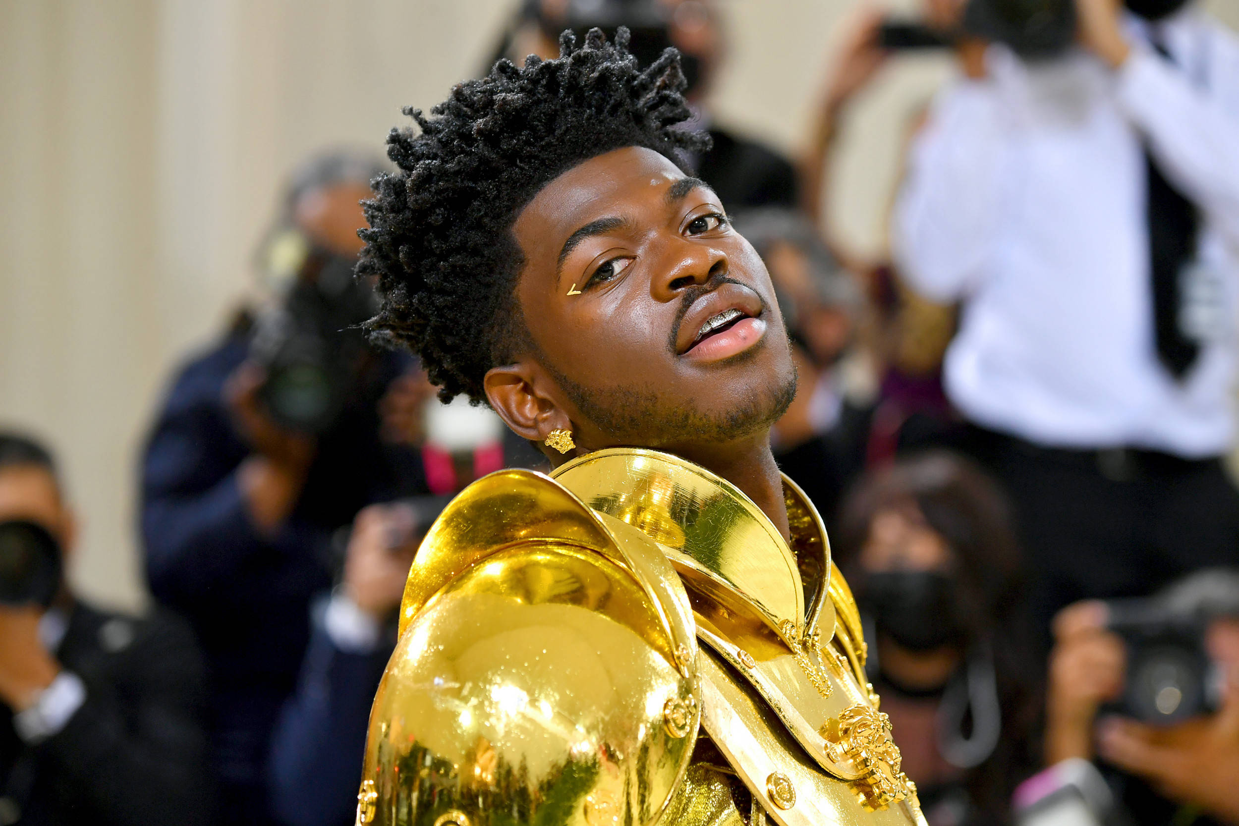 Lil Nas X 金甲裝扮 壁紙 — 一位捲髮年輕男子穿著閃亮金色服裝，在擠滿人的房間裡抬頭看向攝影師。
