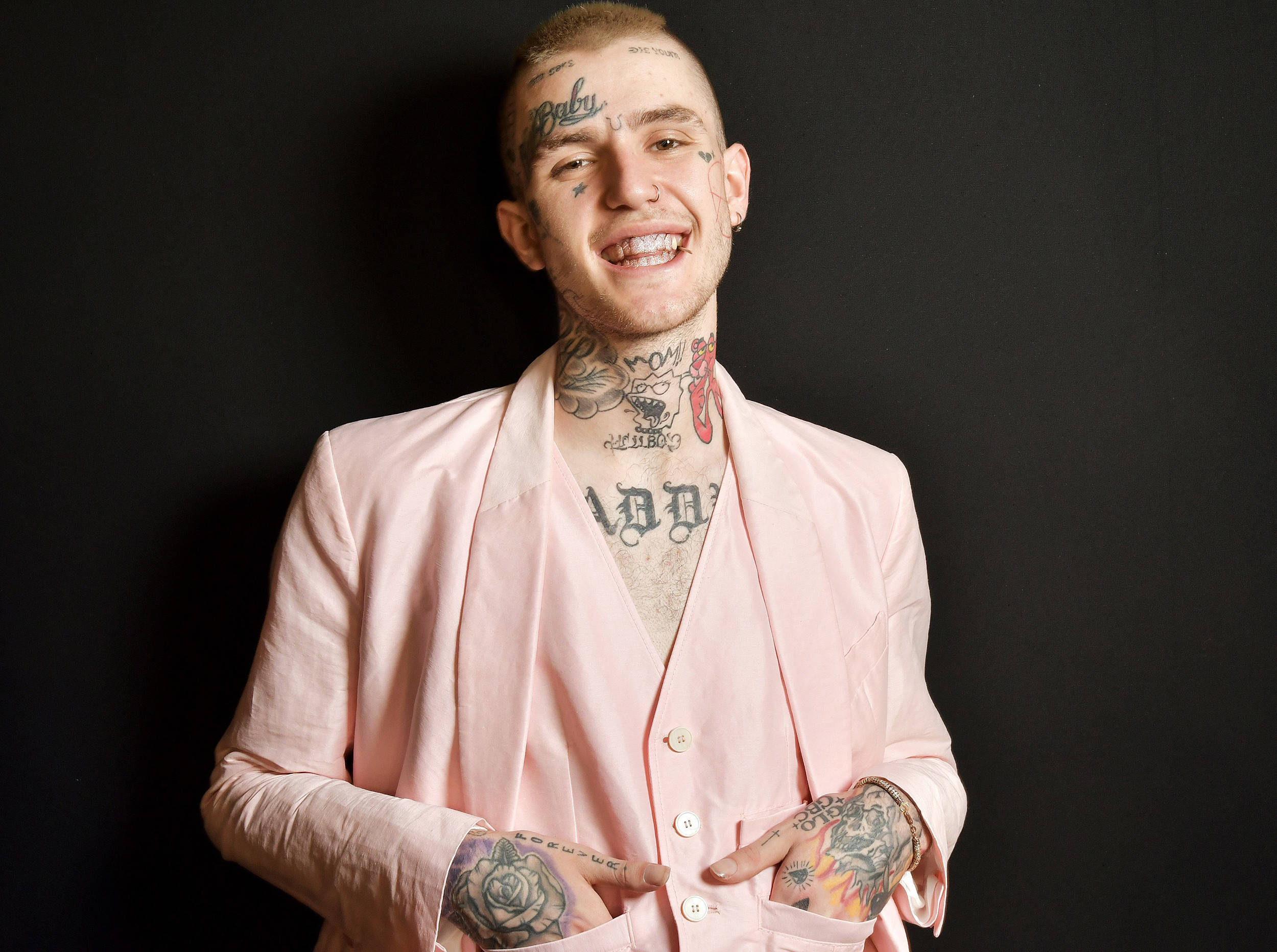 Lil Peep 身穿粉紅西裝登台演出 壁紙 — 一個微笑的男人，身上有大量紋身，穿著粉紅色西裝，背景為黑色。
