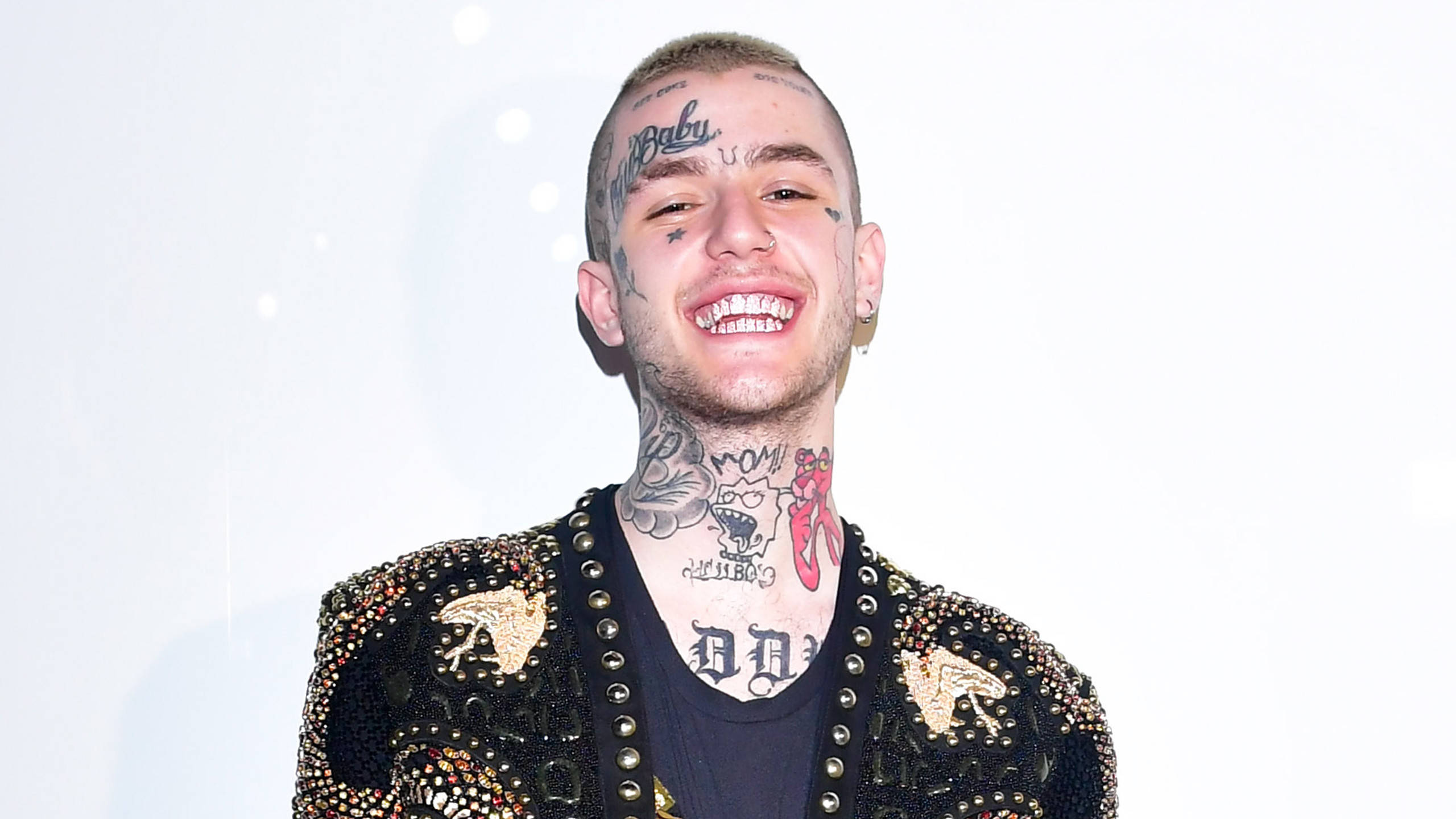 Lil Peep 展現時尚造型 壁紙 — 一個微笑的男人，臉上和脖子上有紋身，穿著一件帶有鉚釘的黑色外套，背景是明亮的白色。