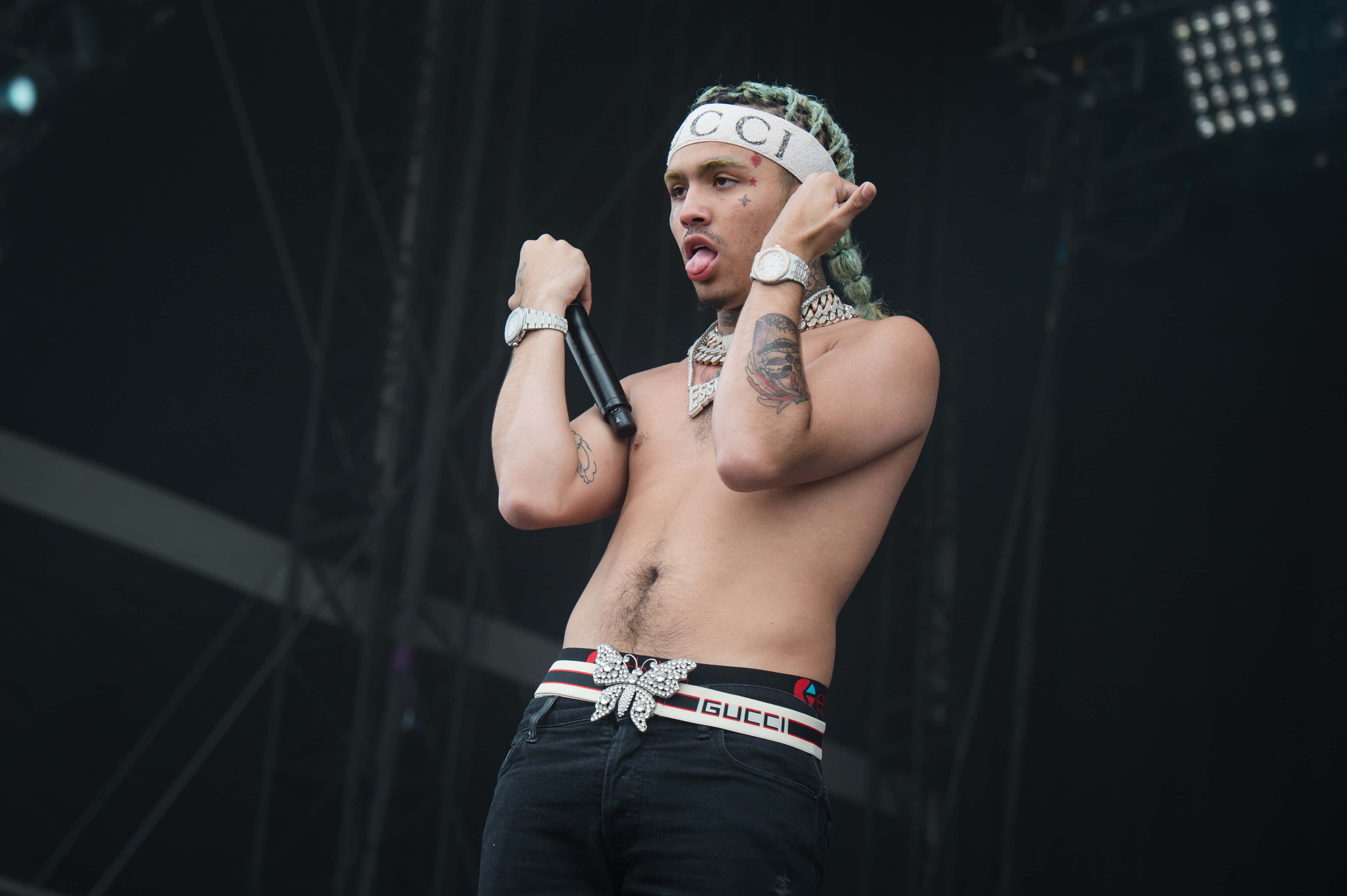 Lil Pump駕駛豪華跑車 壁紙 — 一位赤裸上身的男性表演者，綠色辮子頭髮和Gucci腰帶，在黑暗舞台上明亮燈光下對著麥克風唱歌。
