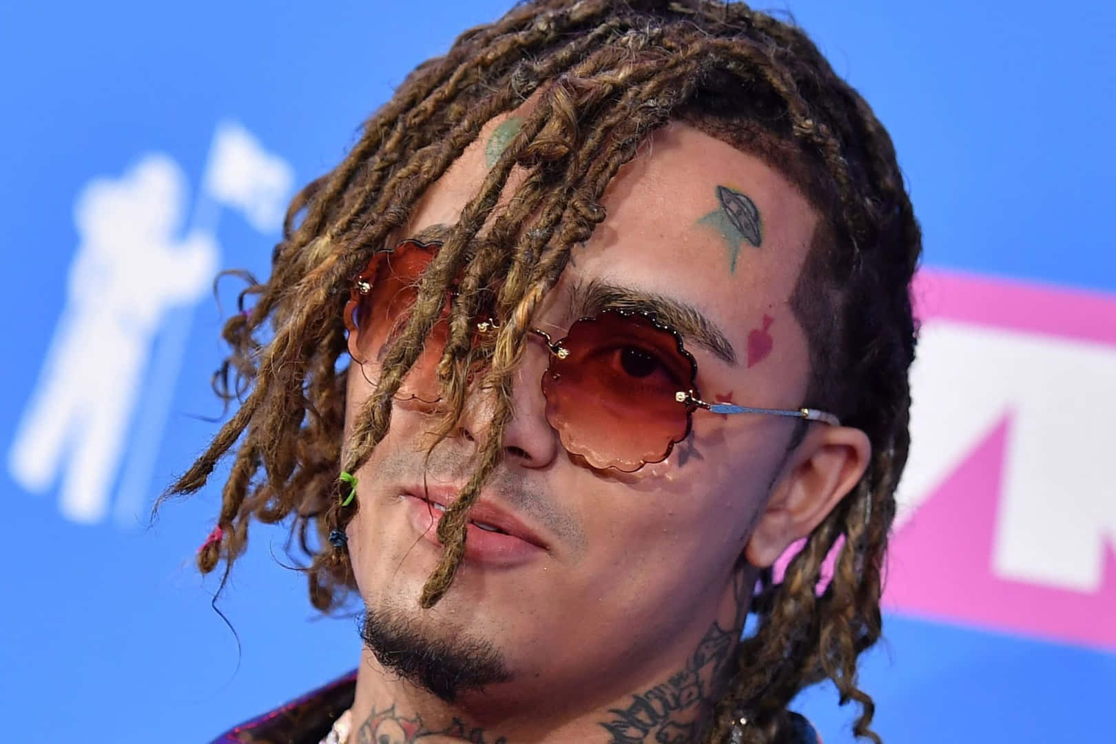 Lil Pump 穿著橙色連帽衫 壁紙 — 一名留著雷鬼辮、有紋身的男子戴著圓形太陽眼鏡，背景為藍色和粉紅色，特寫鏡頭。