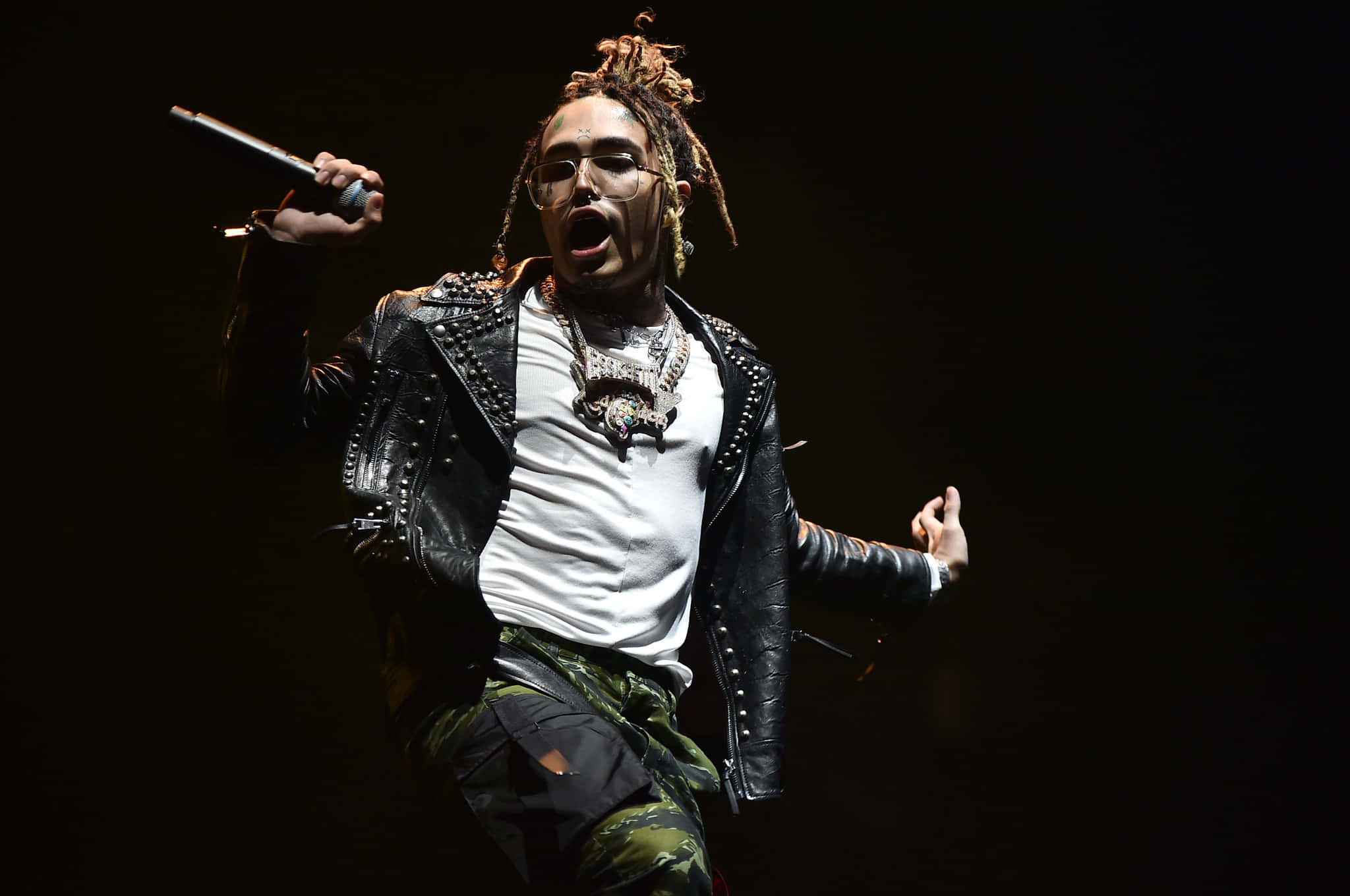 Lil Pump 帶動嘻哈潮流 壁紙 — 一位梳著辮子頭、戴眼鏡的饒舌歌手，身穿飾有鉚釘的皮夾克和迷彩褲，在戲劇性的聚光燈下熱情地在舞台上表演。