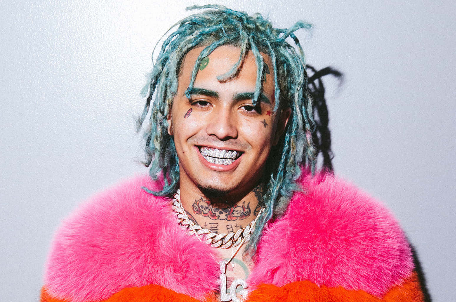說唱歌手 Lil Pump 在紅燈下演出 壁紙 — 一位微笑的男子，留著藍色辮子並有紋身，穿著亮粉紅色毛皮大衣，背景為純灰色。