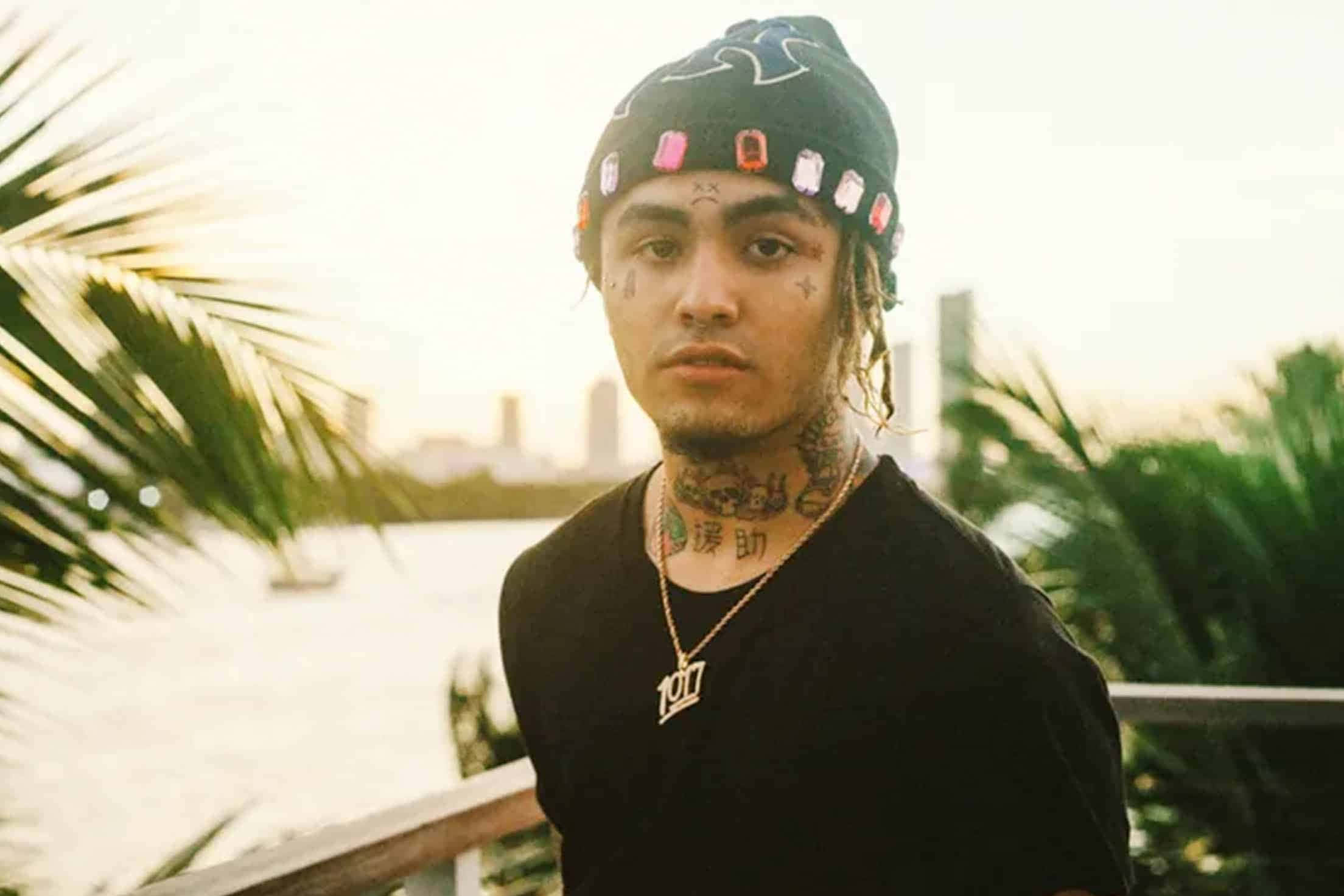 嘻哈天王Lil Pump自信滿溢 壁紙 — 一位年輕男子帶著紋身和冷帽，站在水邊，背景有棕櫚樹和城市天際線，沐浴在柔和的金色光線中。