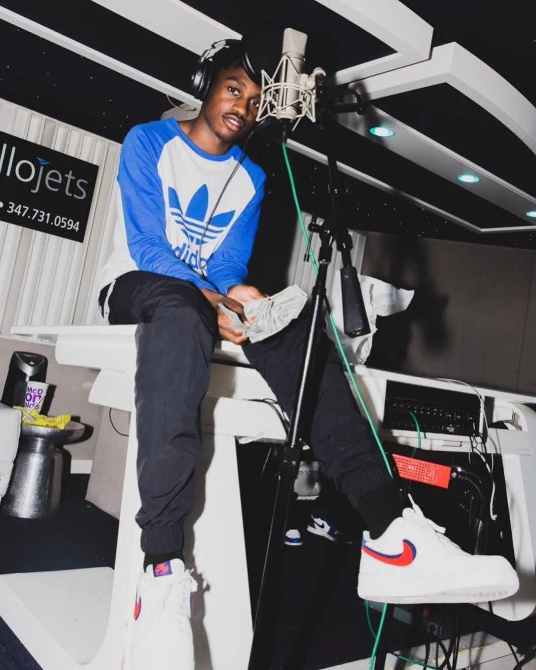 Lil Tjay 在錄音室 壁紙 — 一位身穿藍白Adidas襯衫的年輕男子隨意地坐在白色工作室桌上，手持現金，戴著耳機，靠近麥克風。