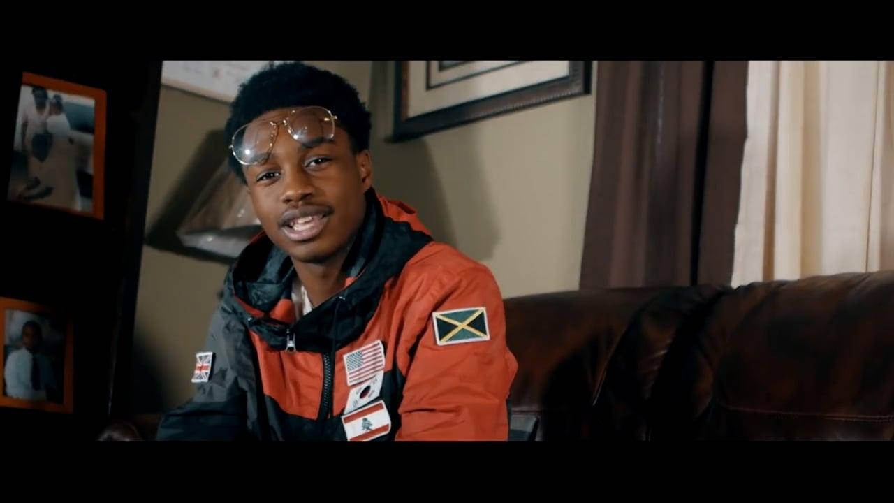Lil Tjay 的歌曲《Resume》 壁紙 — 一名身穿橙色和黑色夾克、帶有旗幟的年輕男子坐在皮革沙發上，房間光線溫暖，牆上掛著裱框照片。