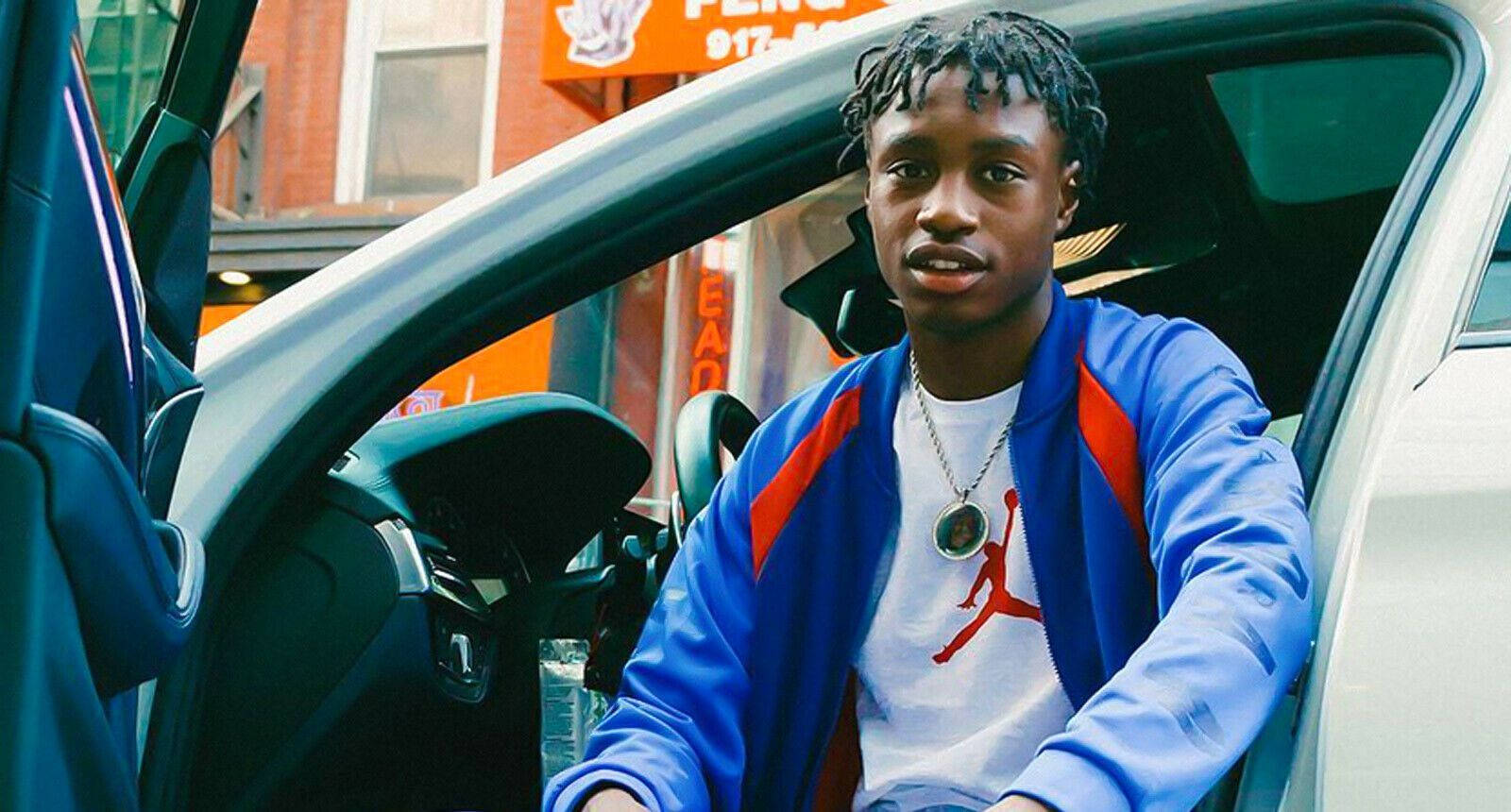 Lil Tjay 車內拍攝照 壁紙 — 一名身穿藍色外套和Jordan T恤的年輕男子坐在車內，車門敞開，背景是城市風光。