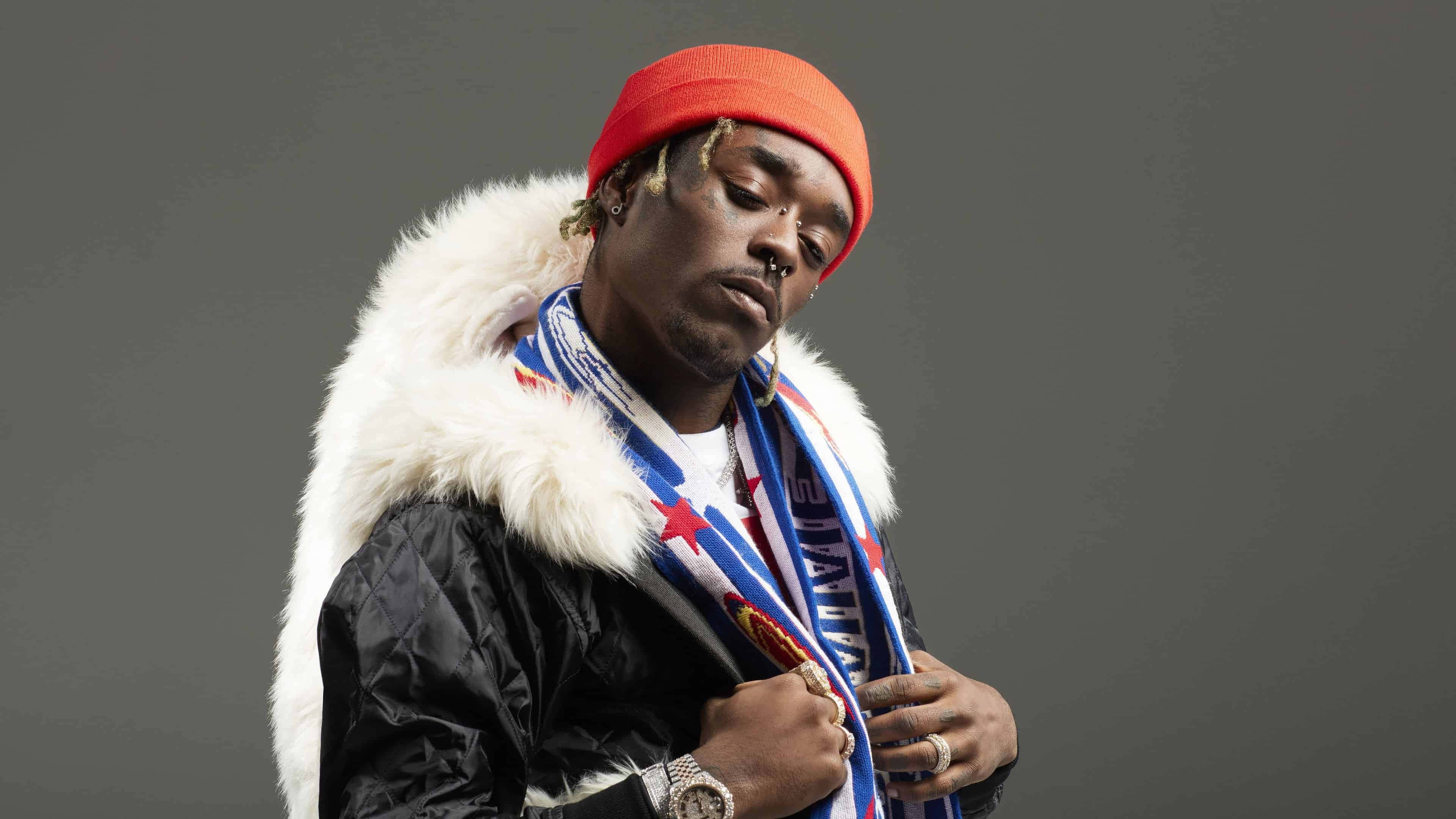 Lil Uzi Vert 4K 桌面壁紙 壁紙 — 一個戴紅色冷帽、穿毛領外套的男人，在純灰色背景前向下望。
