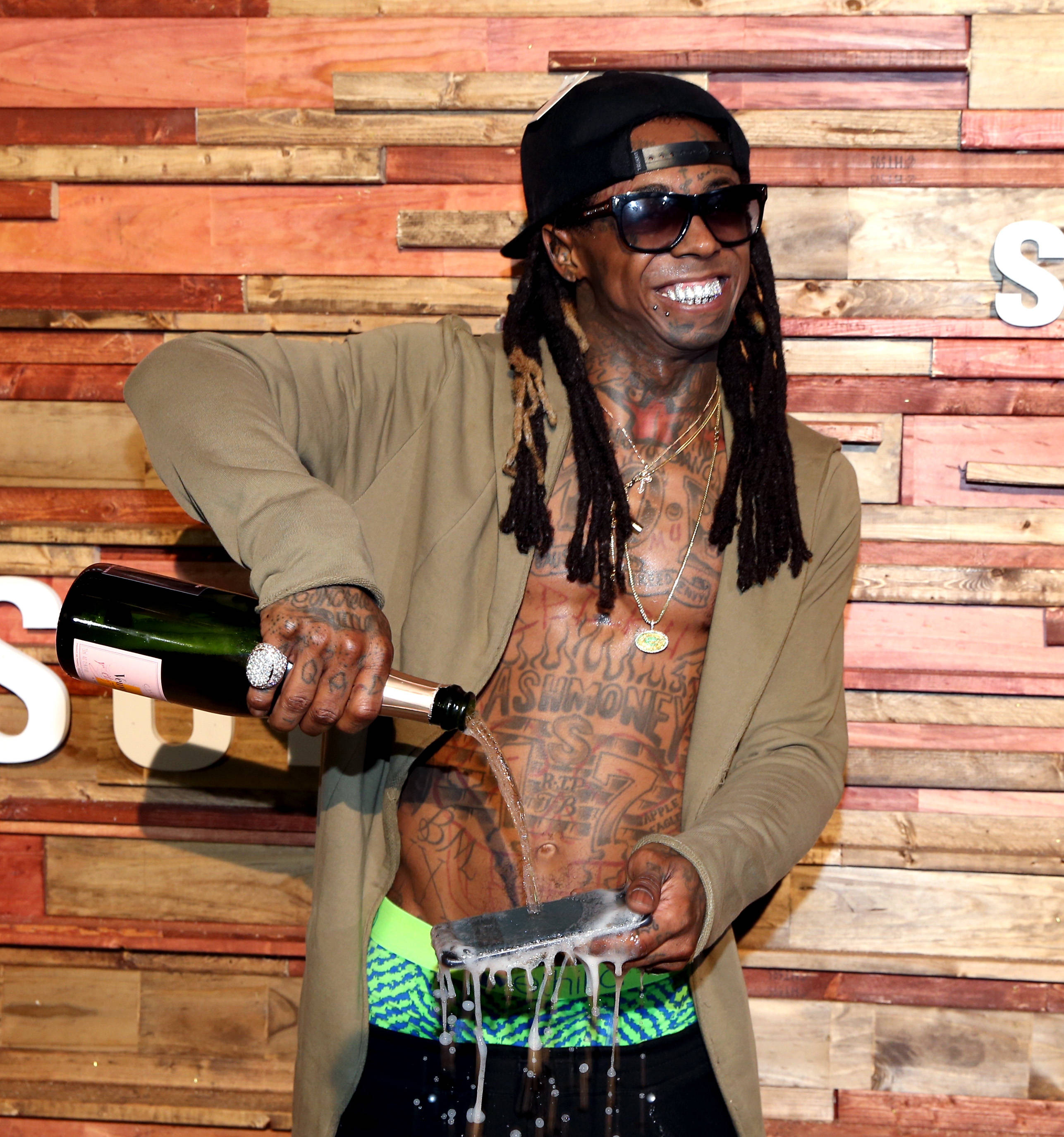 Lil Wayne 倒香檳 壁紙 — 一個有紋身的男人戴著黑色帽和太陽眼鏡，在木牆背景下將香檳倒在金屬托盤上。