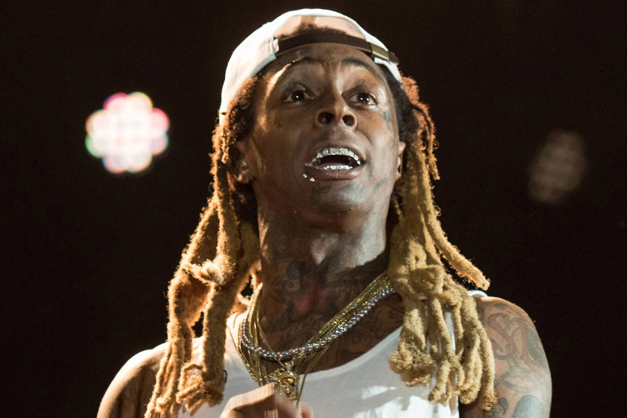 Lil Wayne 內搭背心 壁紙 — 一位帶有紋身、梳著辮子頭並戴著白色帽子的饒舌歌手在舞台燈光下表演的特寫。