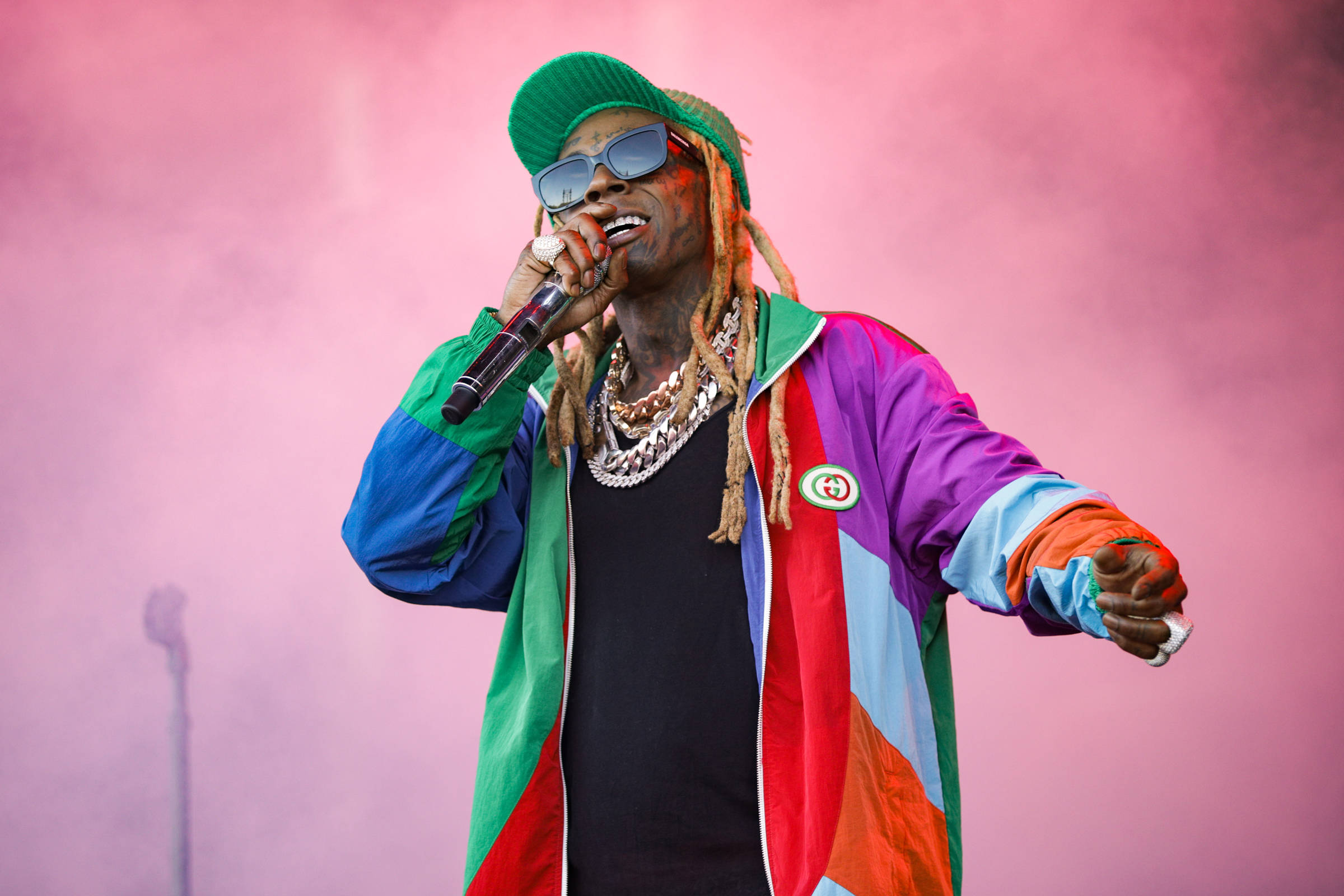 Lil Wayne 唱歌 壁紙 — 一位穿著彩色外套和綠色冷帽的饒舌歌手，在模糊的粉紅色煙霧背景下對著咪高峰演唱。