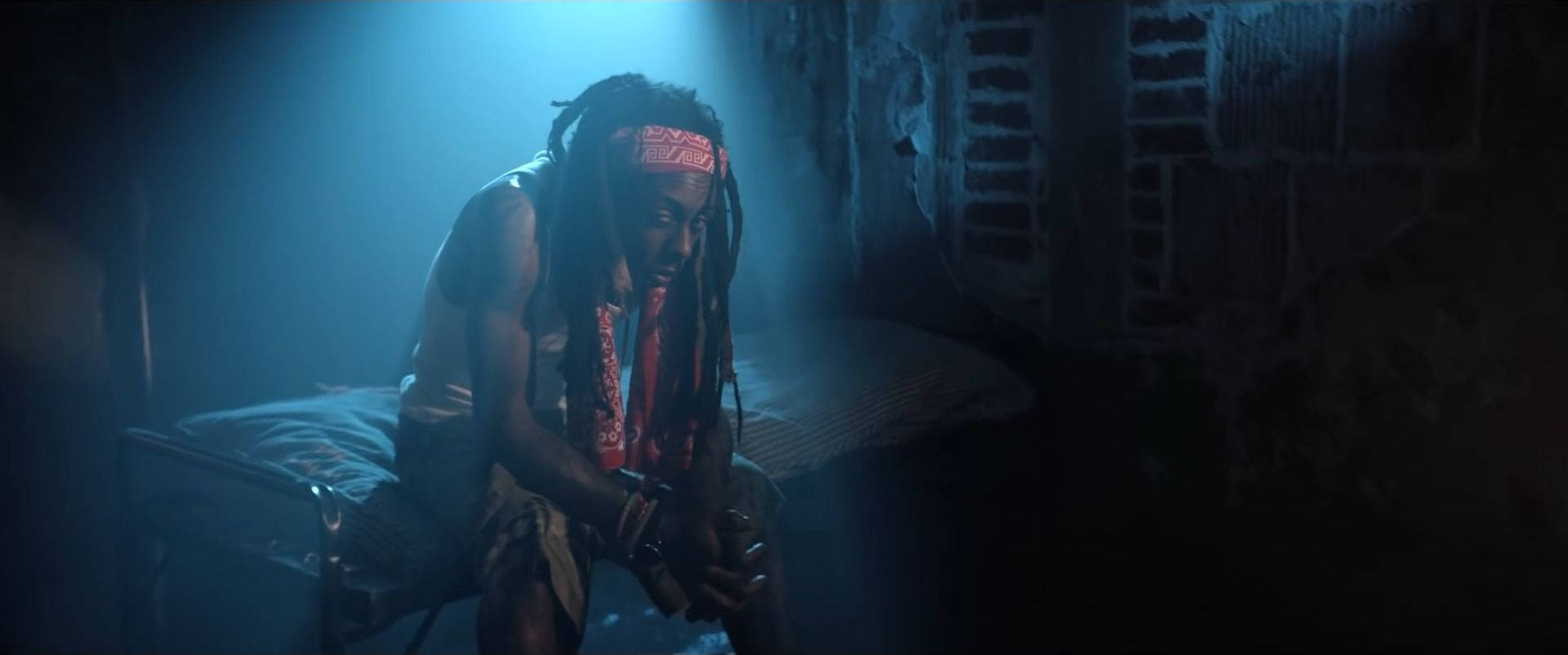 Lil Wayne — 嘻哈樂壇的傳奇天才 壁紙 — 一名留著辮子頭的男人蜷縮著坐在床上，房間光線昏暗，藍色燈光，牆壁剝落，百葉窗破舊。