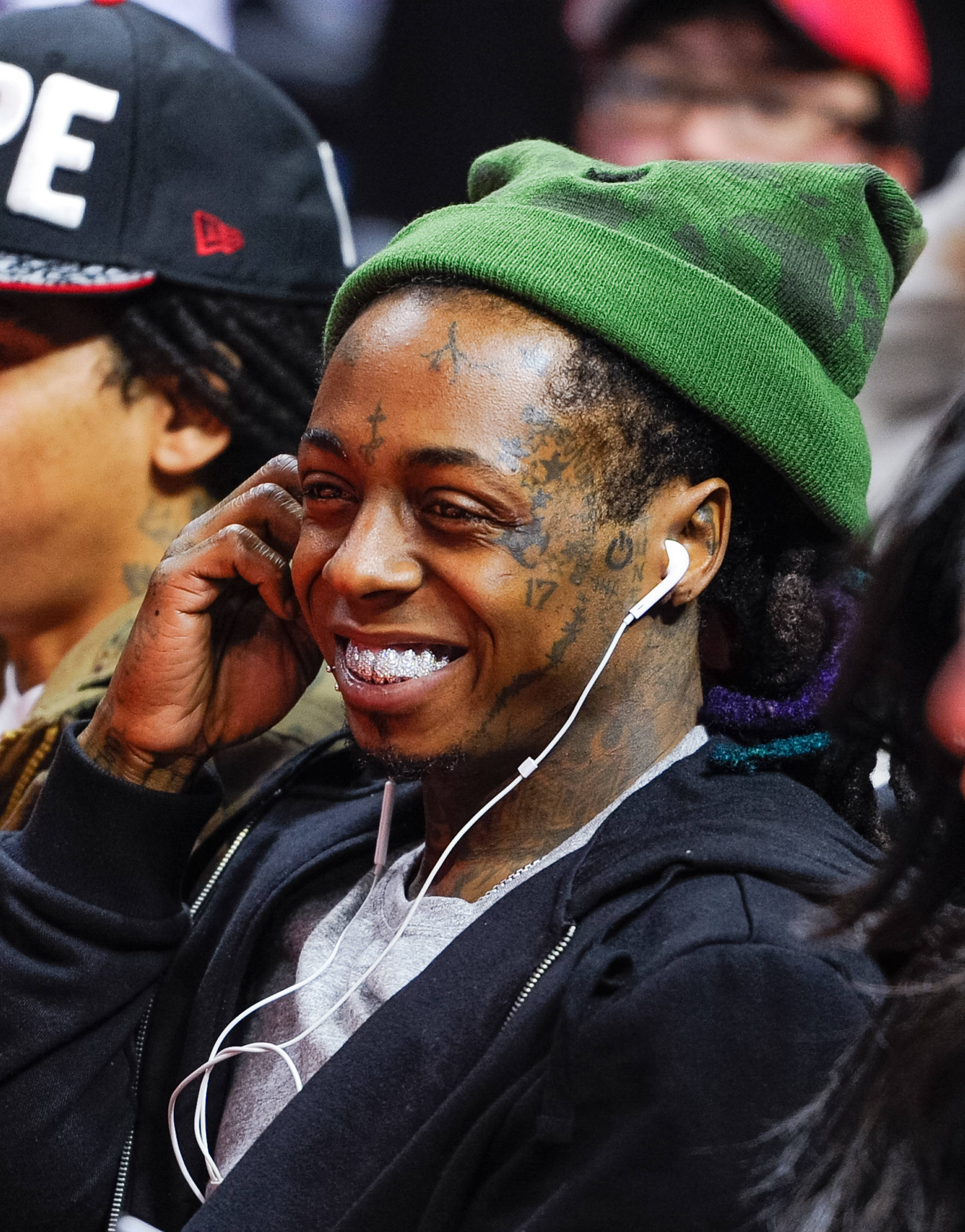 Lil Wayne 大笑 壁紙 — 一個面帶微笑、有紋身、戴綠色冷帽嘅男人喺擠迫嘅活動中用白色耳塞聽音樂。