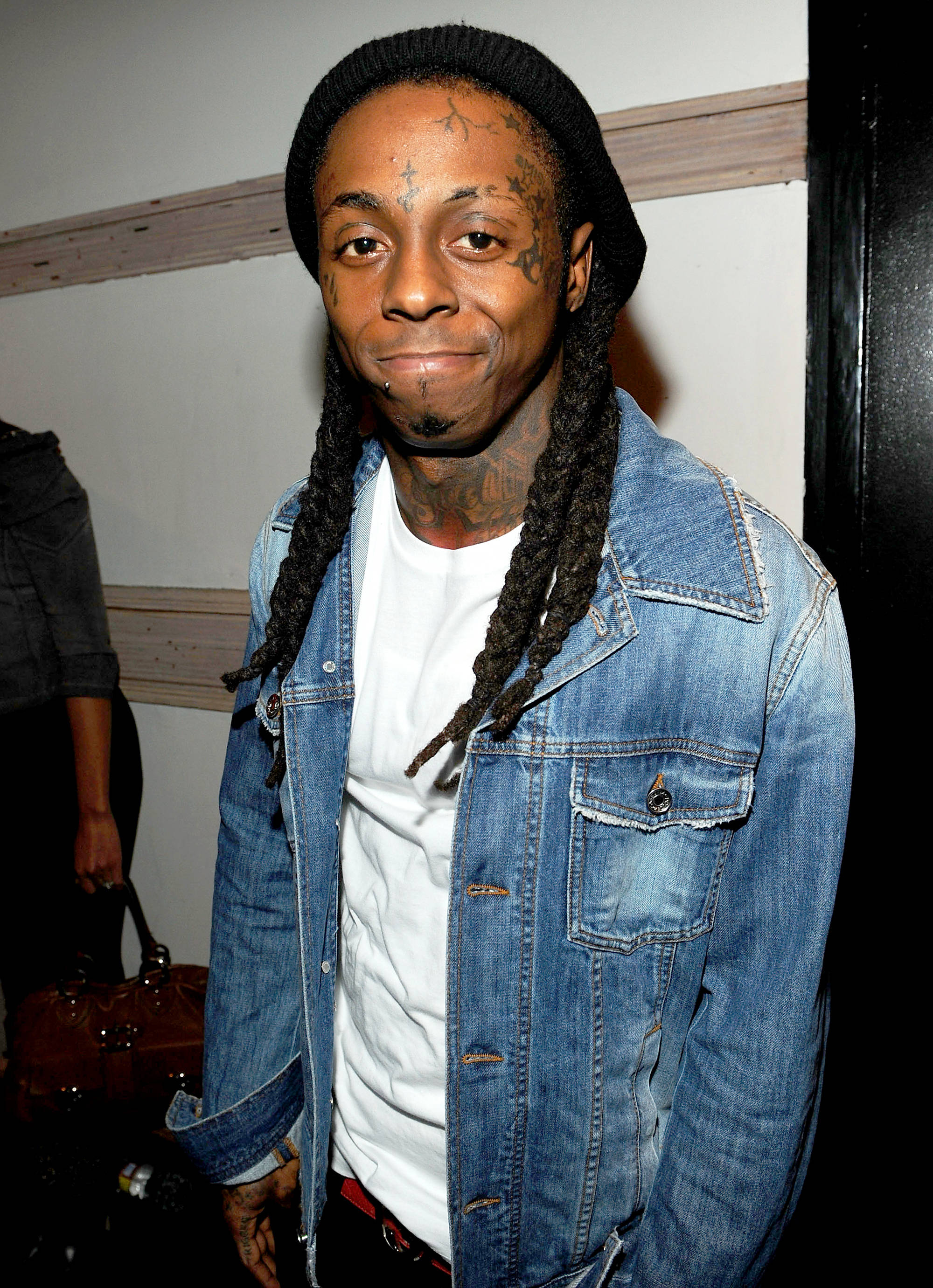 Lil Wayne 微笑 壁紙 — 一名留著辮子頭和紋身的男子，在室內人造燈光下，身穿牛仔外套內搭白色T恤。