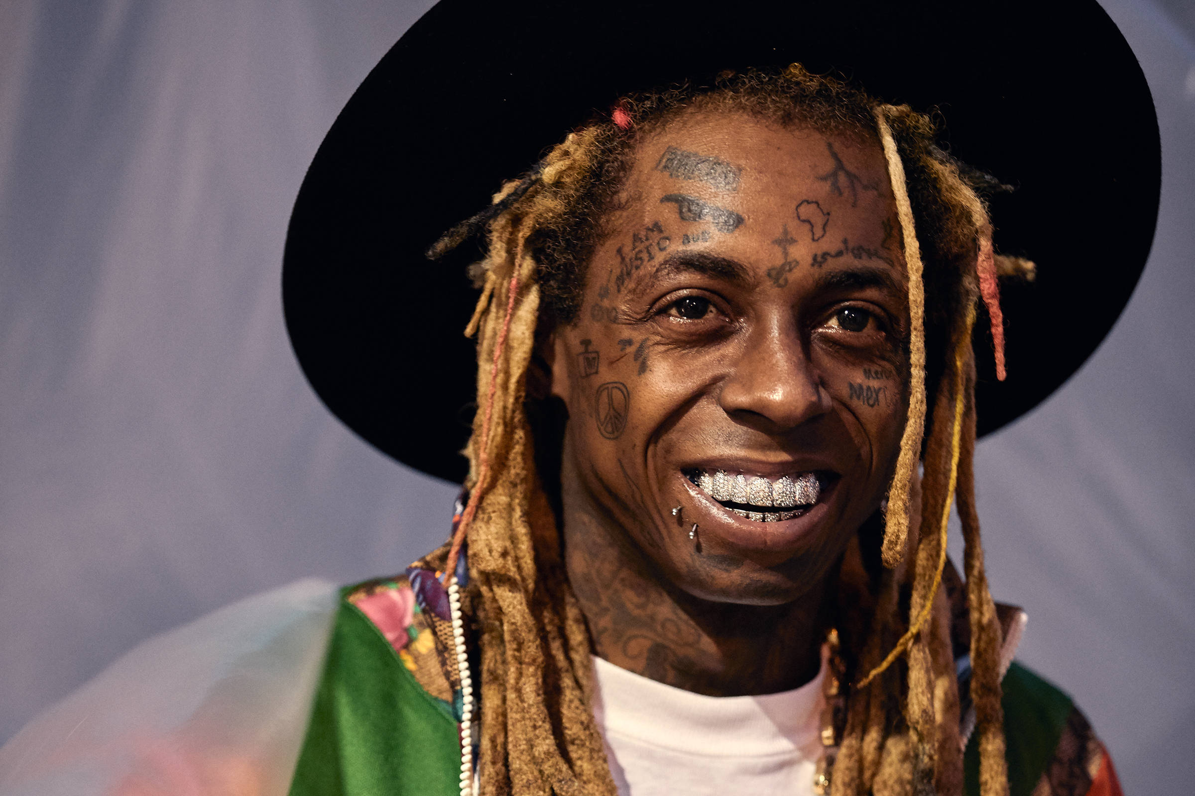Lil Wayne 戴大帽子 壁紙 — 一個微笑的男人，臉上紋身，留著辮子，戴著黑色帽子和彩色外套，背景樸素。
