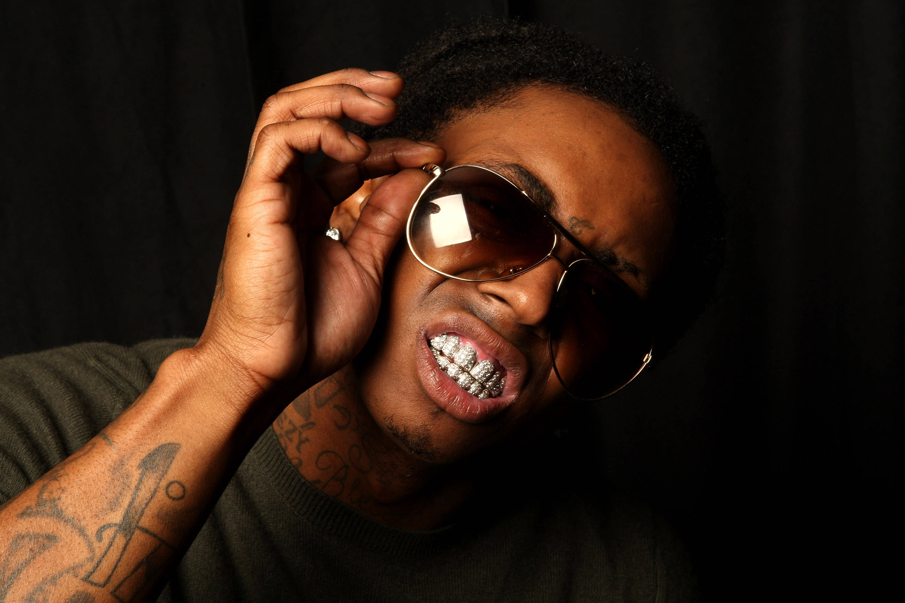 Lil Wayne 戴太陽眼鏡 壁紙 — 一名有紋身和金色牙套的男子在黑色背景下調整太陽眼鏡。