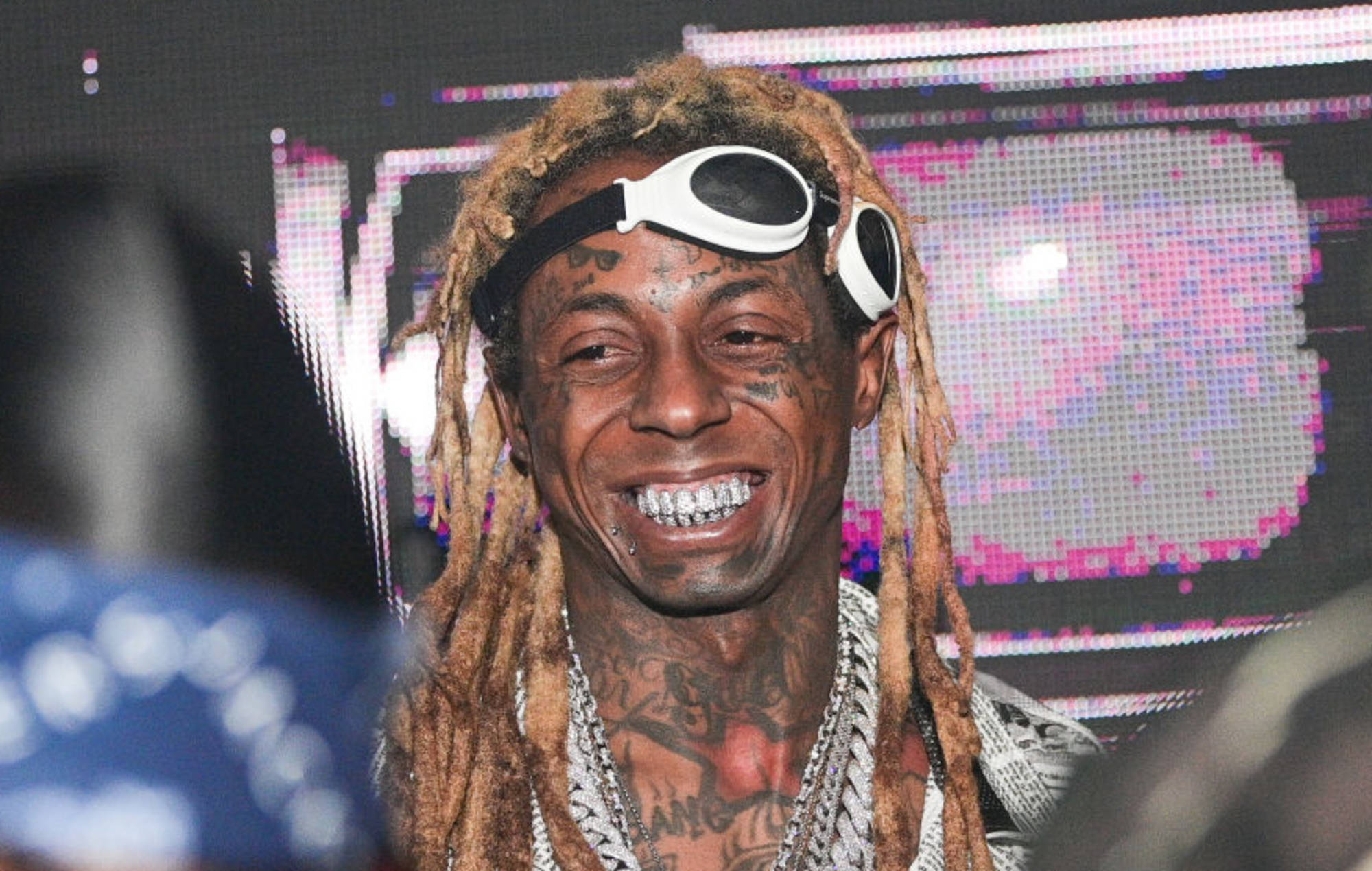 Lil Wayne 戴眼鏡 壁紙 — 一個微笑的男人，留著辮子頭和護目鏡，在發光的像素背景前對著鏡頭咧嘴笑。