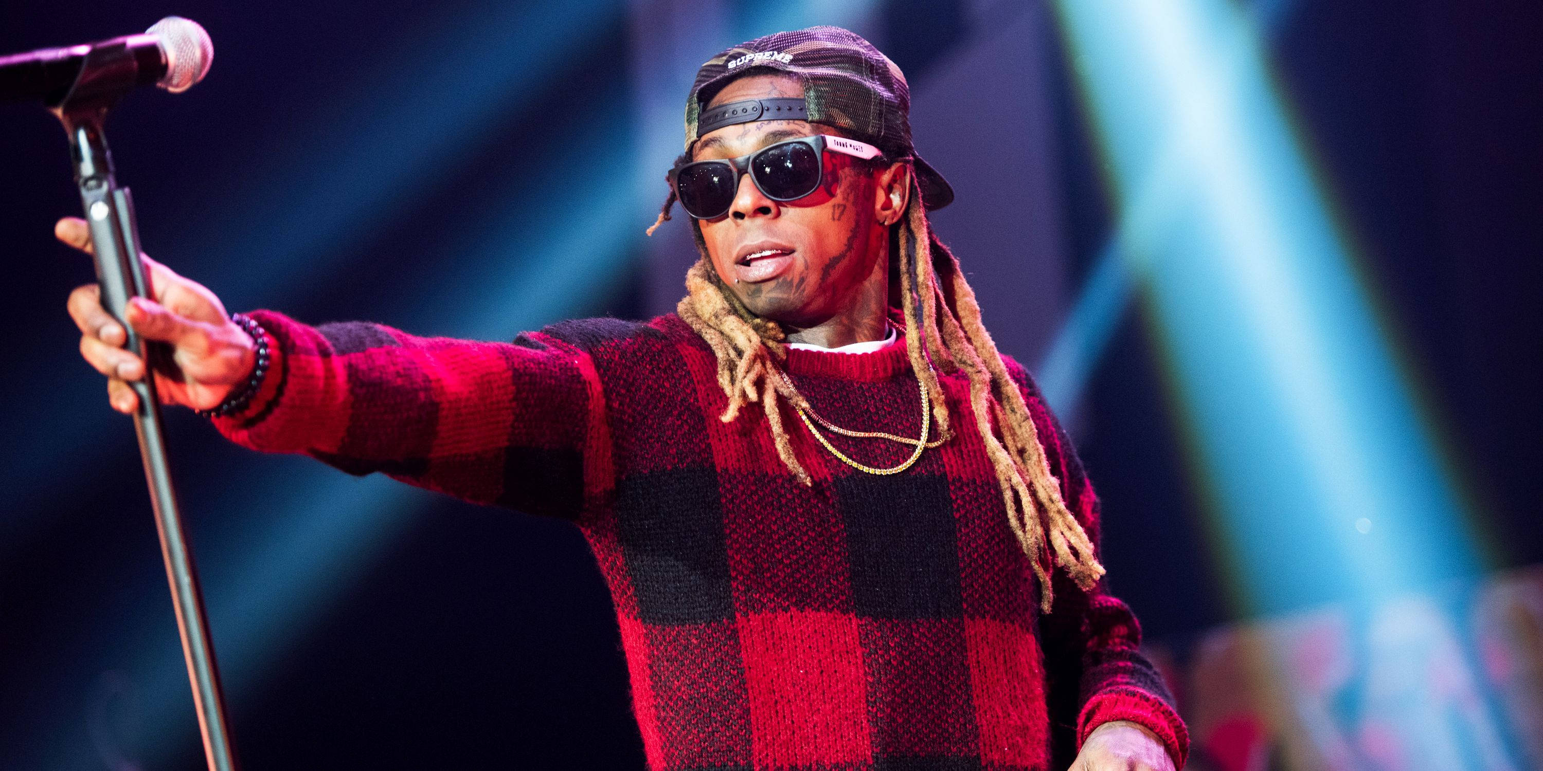 Lil Wayne 拿麥克風架 壁紙 — 一位穿著紅色格仔毛衣和太陽眼鏡的饒舌歌手在藍色聚光燈下拿著麥克風在舞台上表演。