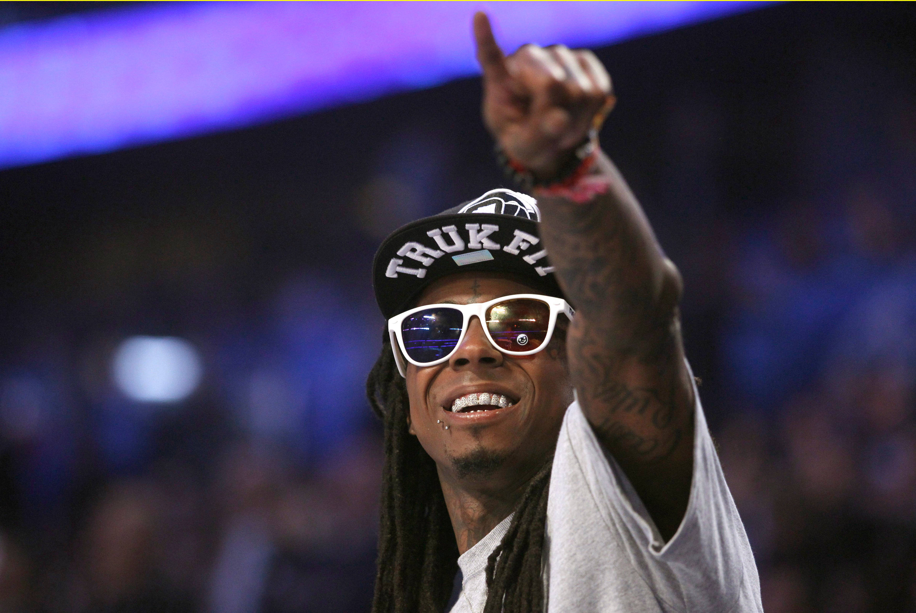 Lil Wayne 指著前方 壁紙 — 一個微笑的男人，梳著辮子頭，戴著白色太陽眼鏡，在紫色舞台燈光下的演唱會中向上指。