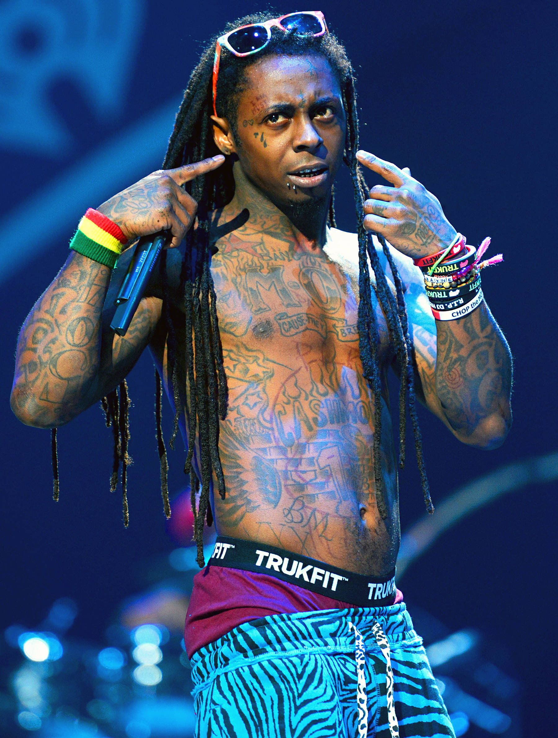 Lil Wayne 穿短褲 壁紙 — 一位身上有紋身、頭戴太陽眼鏡、留著雷鬼辮的音樂家在昏暗的舞台上指著自己的太陽穴。