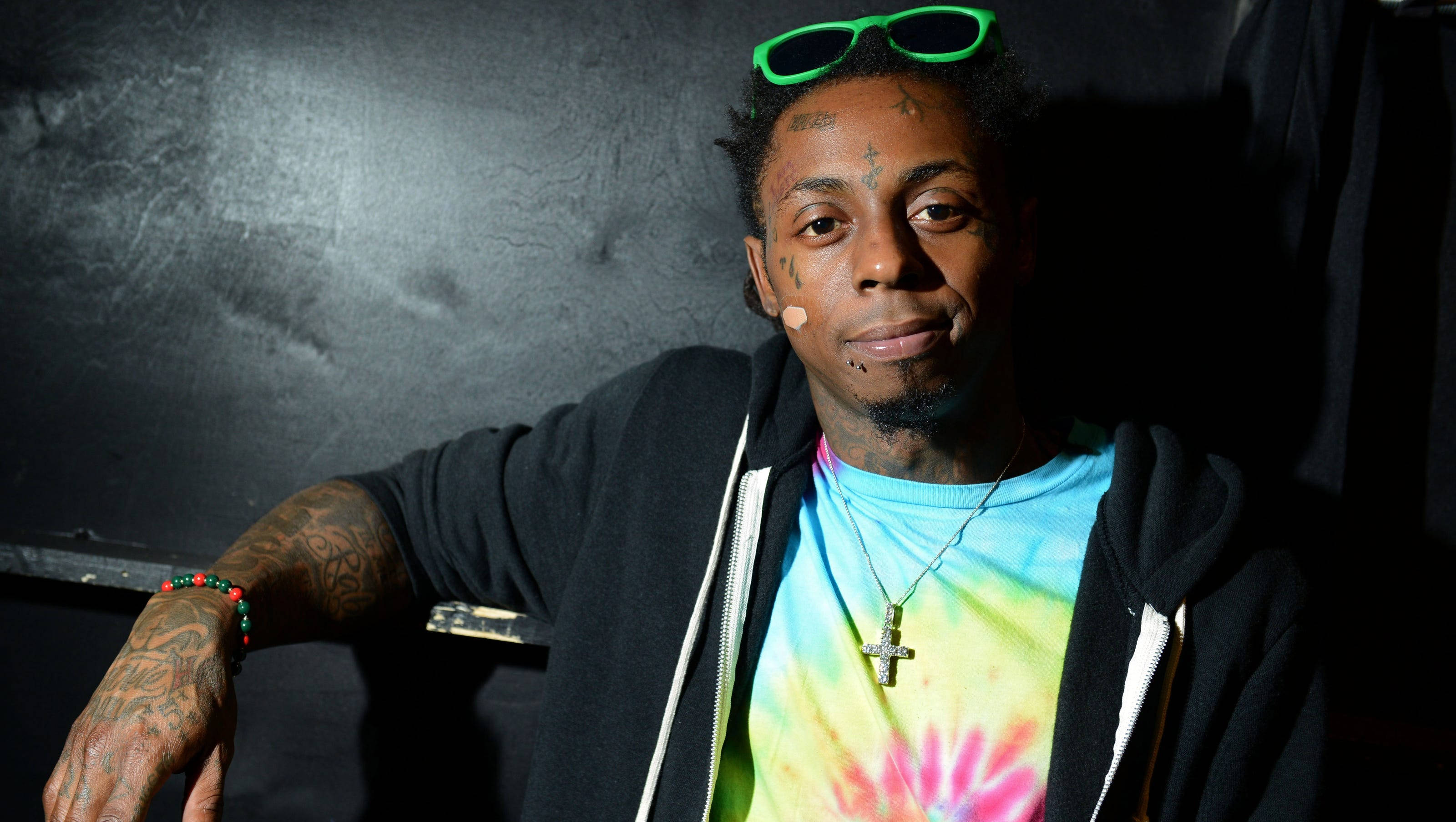 Lil Wayne 簡約微笑 壁紙 — 一個有紋身、頭上戴著綠色太陽眼鏡的男人，在深色紋理牆壁前穿著黑色連帽衫下的紮染襯衫。