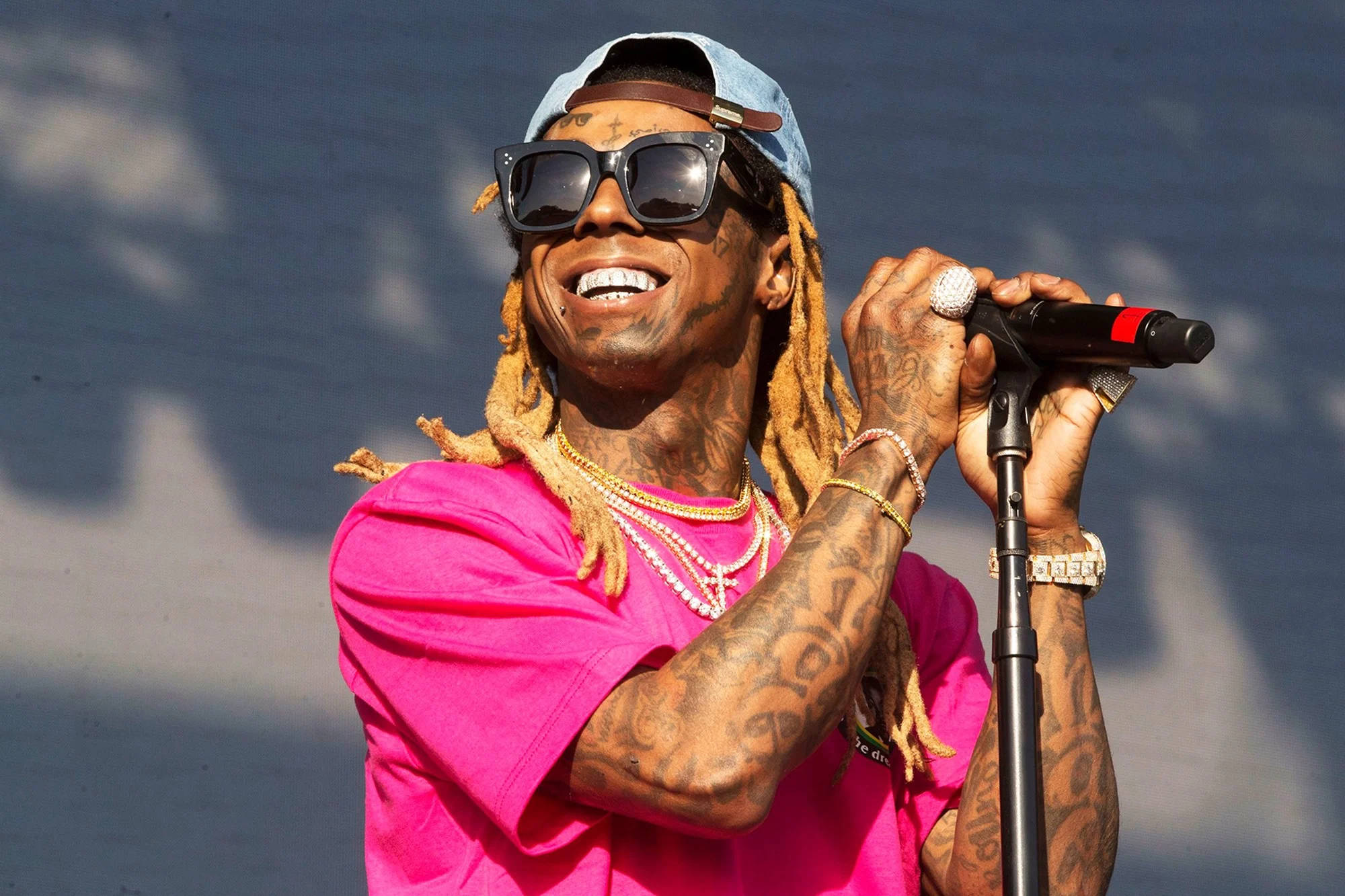 Lil Wayne 粉色襯衫 壁紙 — 一位微笑的饒舌歌手，留著辮子頭和刺青，穿著亮粉色襯衫和太陽眼鏡對著麥克風表演。