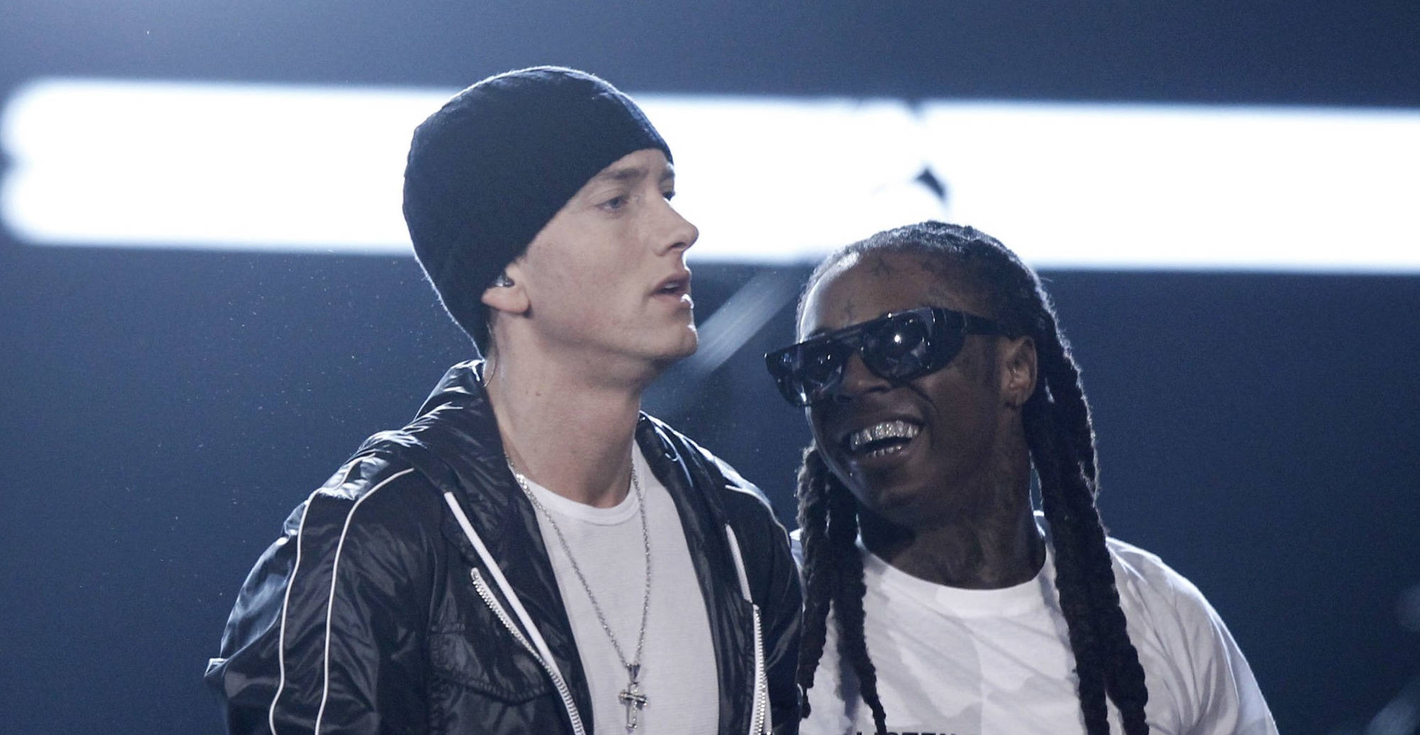 Lil Wayne 與 Eminem 壁紙 — 兩位饒舌歌手在舞台上，一位戴黑色冷帽和外套，另一位戴太陽眼鏡和辮子，兩人在明亮的舞台燈光下微笑。