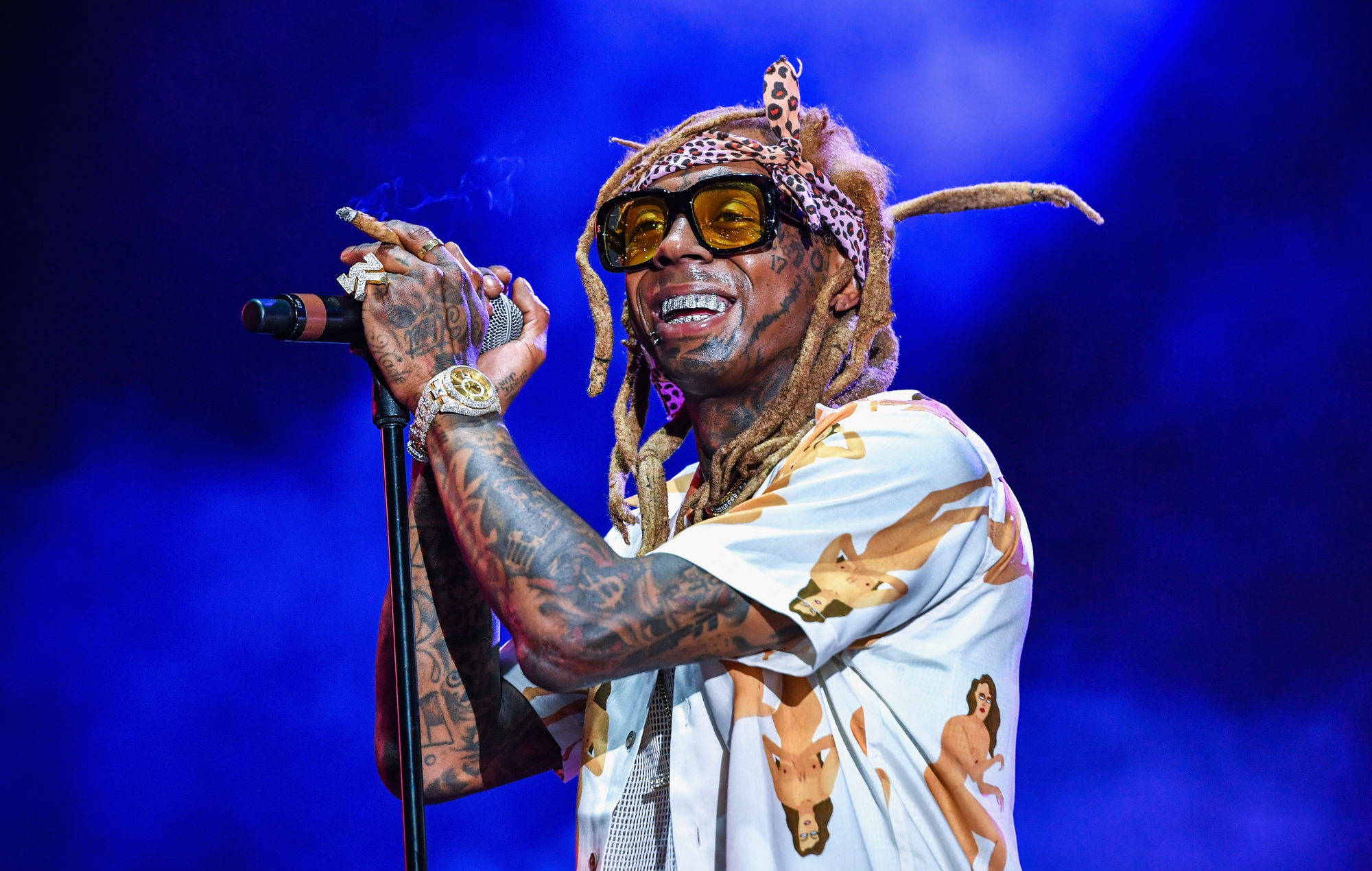 Lil Wayne 藍色背景 壁紙 — Lil Wayne 在舞台上穿著圖案襯衫和太陽眼鏡，手持麥克風，在藍色舞台燈光下吸煙。