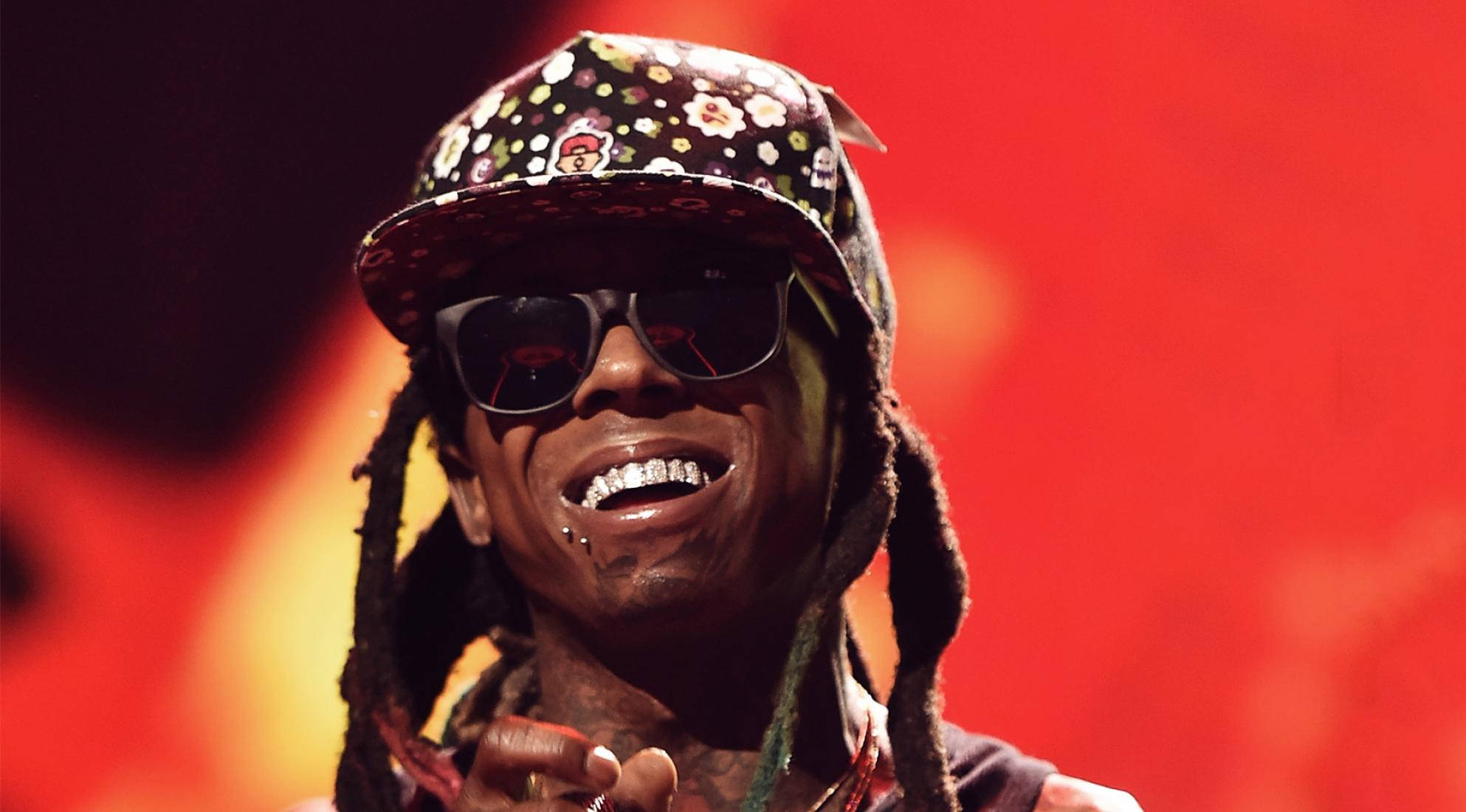 Lil Wayne戴太陽眼鏡 壁紙 — 一位微笑的饒舌歌手，留著辮子頭，戴著花卉圖案帽子，在舞台燈光下對著火紅背景咧嘴笑。