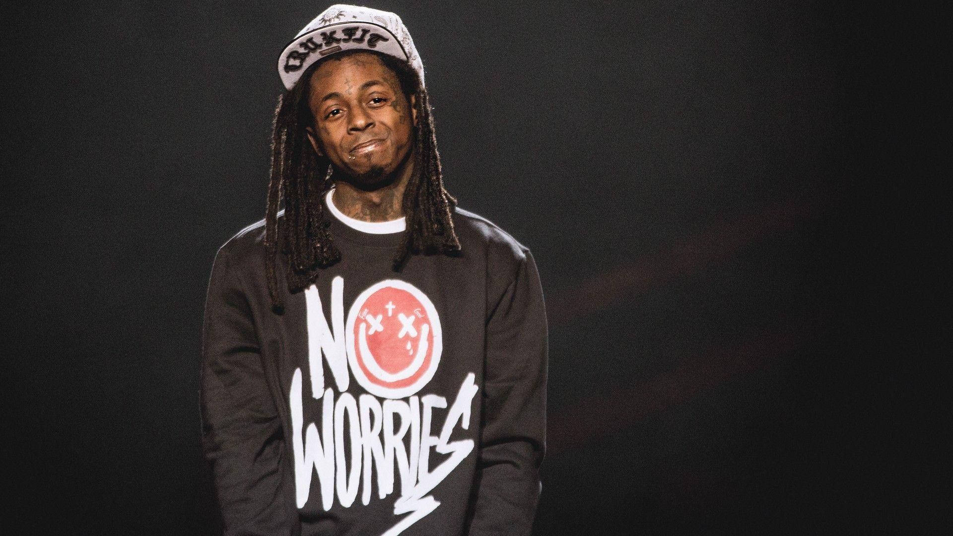 Lil Wayne沉思中 壁紙 — 一名留著辮子、戴著帽子的男士微笑著，身穿黑色衛衣，上面有紅色笑臉和白色「NO WORRIES」字樣。