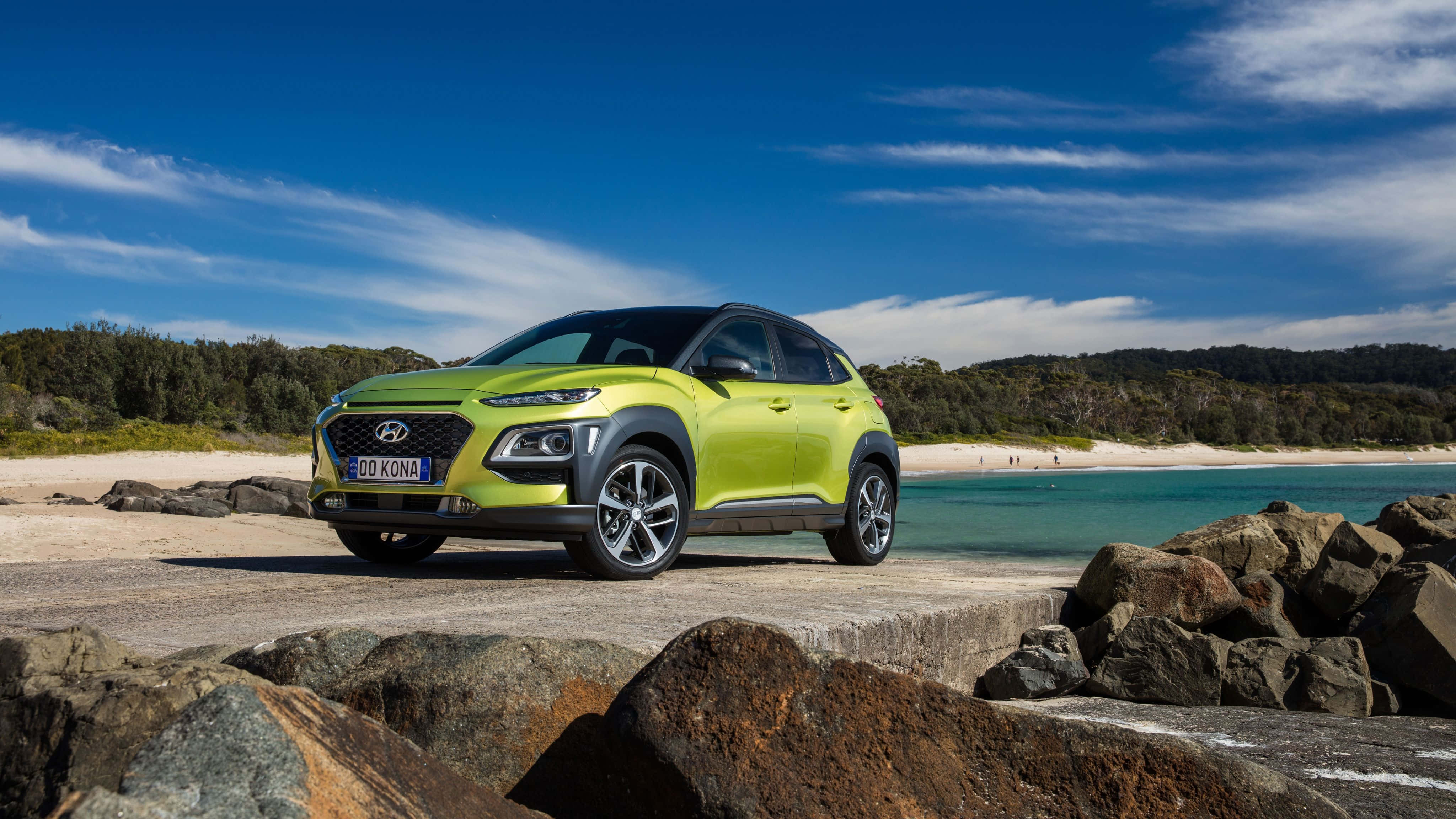 Hyundai Kona 桌布 壁紙 — 一輛充滿活力的青檸綠色現代Kona SUV停泊在岩石海岸小徑旁，旁邊是碧綠的海水，上方是明亮的藍天。