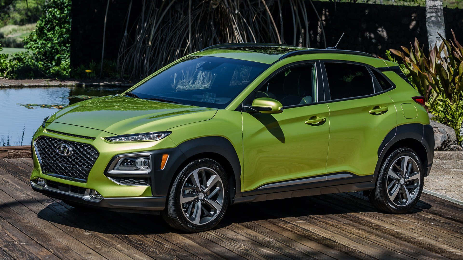 Hyundai Kona 桌布 壁紙 — 一輛充滿活力的青檸綠色現代 Kona SUV 停泊在寧靜池塘旁的木甲板上，周圍有茂密的熱帶植物。