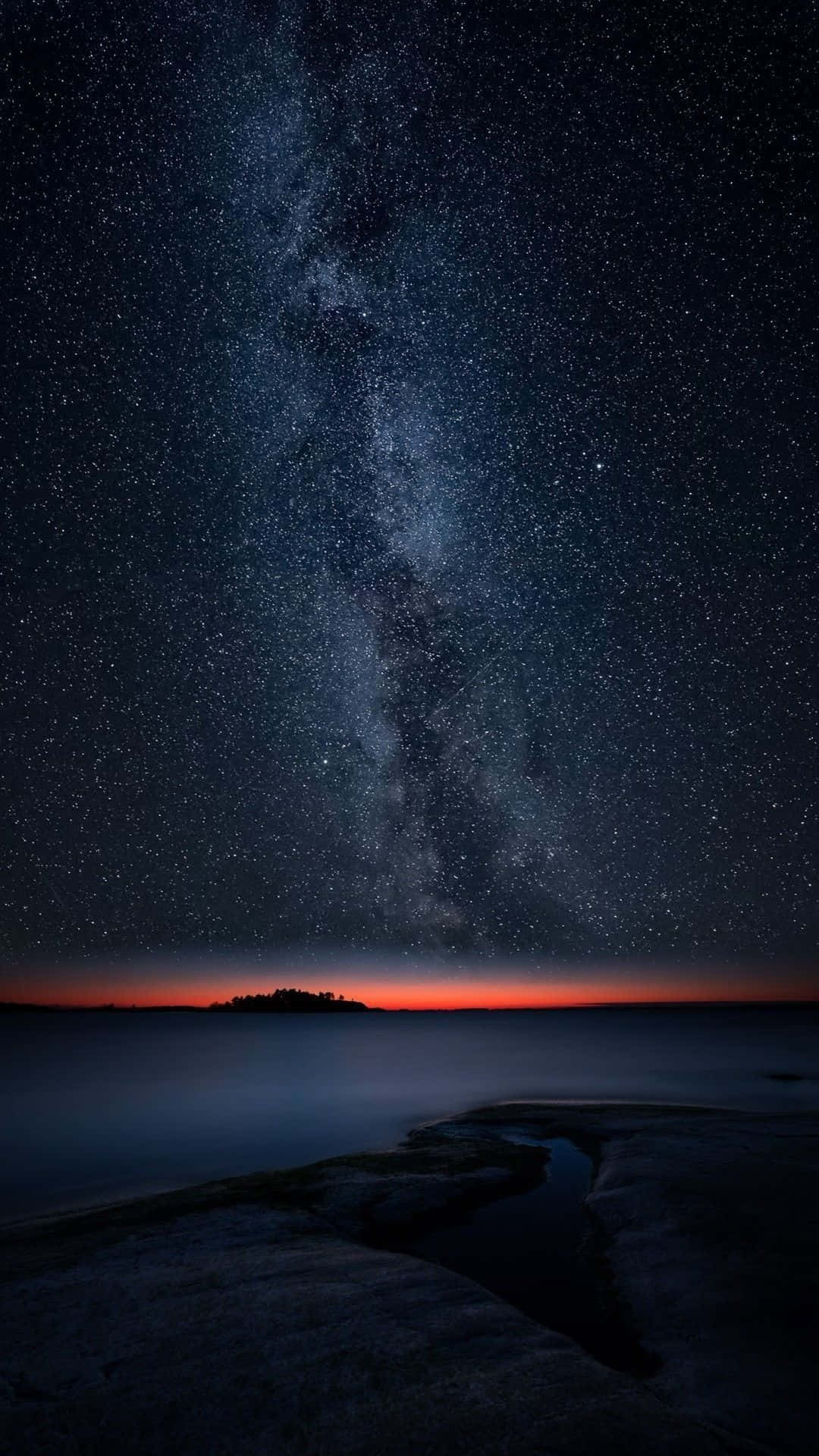  壁紙 — 令人屏息的夜空佈滿繁星，銀河在平靜的海洋上空拱起，遠處有島嶼的輪廓。