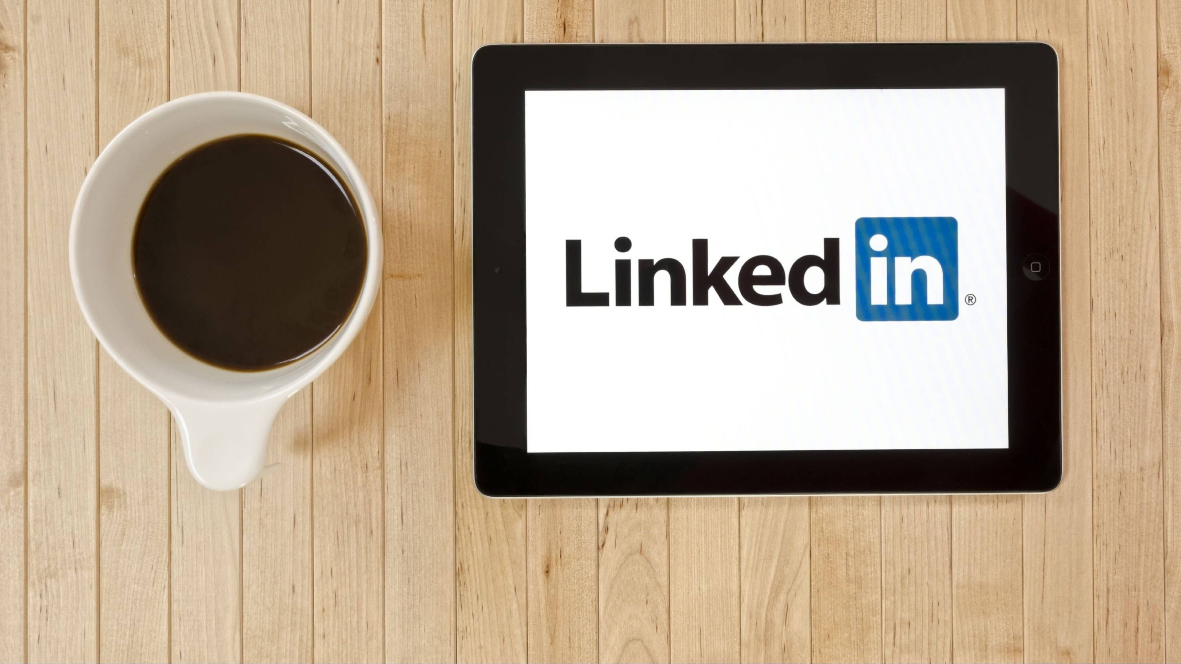 LinkedIn 咖啡平鋪照 壁紙 — 一部顯示LinkedIn標誌的平板電腦放在淺色木桌上，旁邊有一杯白杯黑咖啡。